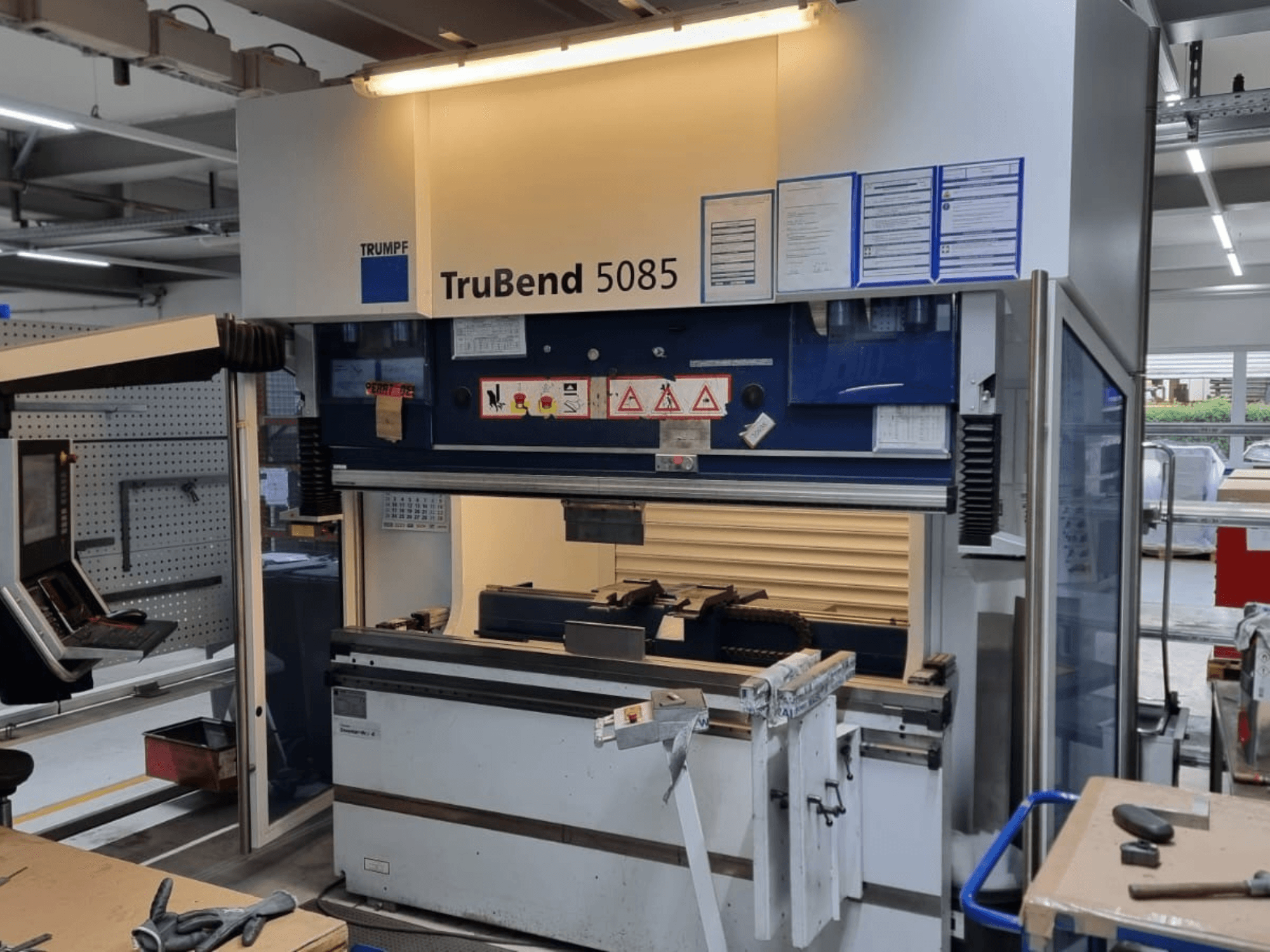 Máquina de dobragem TRUMPF TruBend 5085, vista frontal, mostrando o painel de controlo, estação de operação e avisos de segurança dentro da oficina.