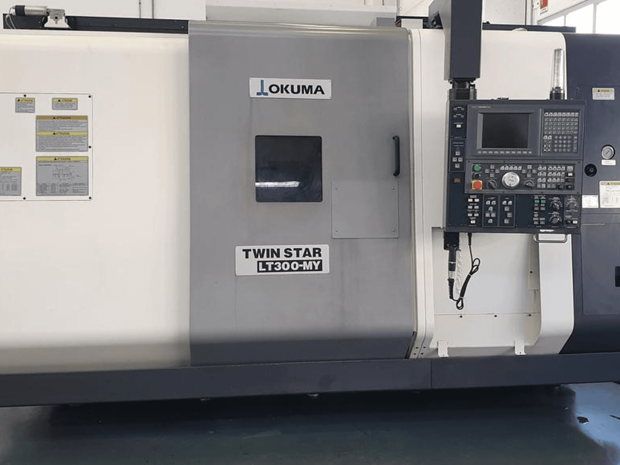 Torno CNC Okuma Twin Star LT300-MY em vista lateral, com painel de controlo, botões operacionais e etiquetas de segurança.