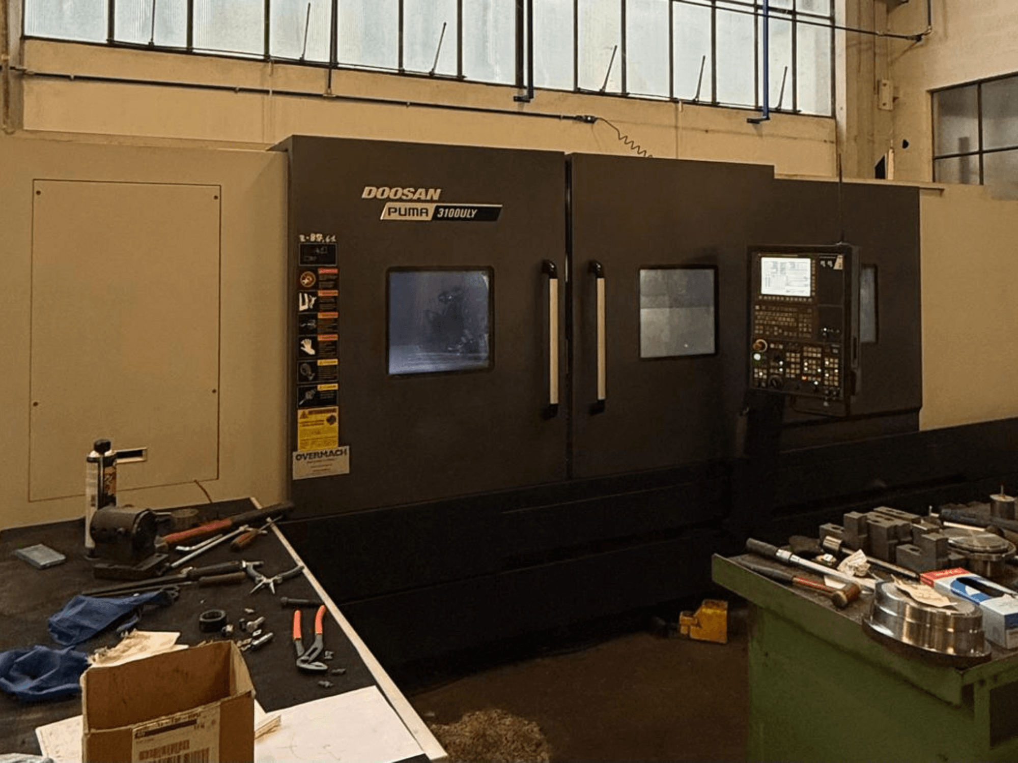 Torno CNC Doosan Puma 3100LY numa oficina, vista lateral, com painel de controlo e ferramentas visíveis na bancada de trabalho.
