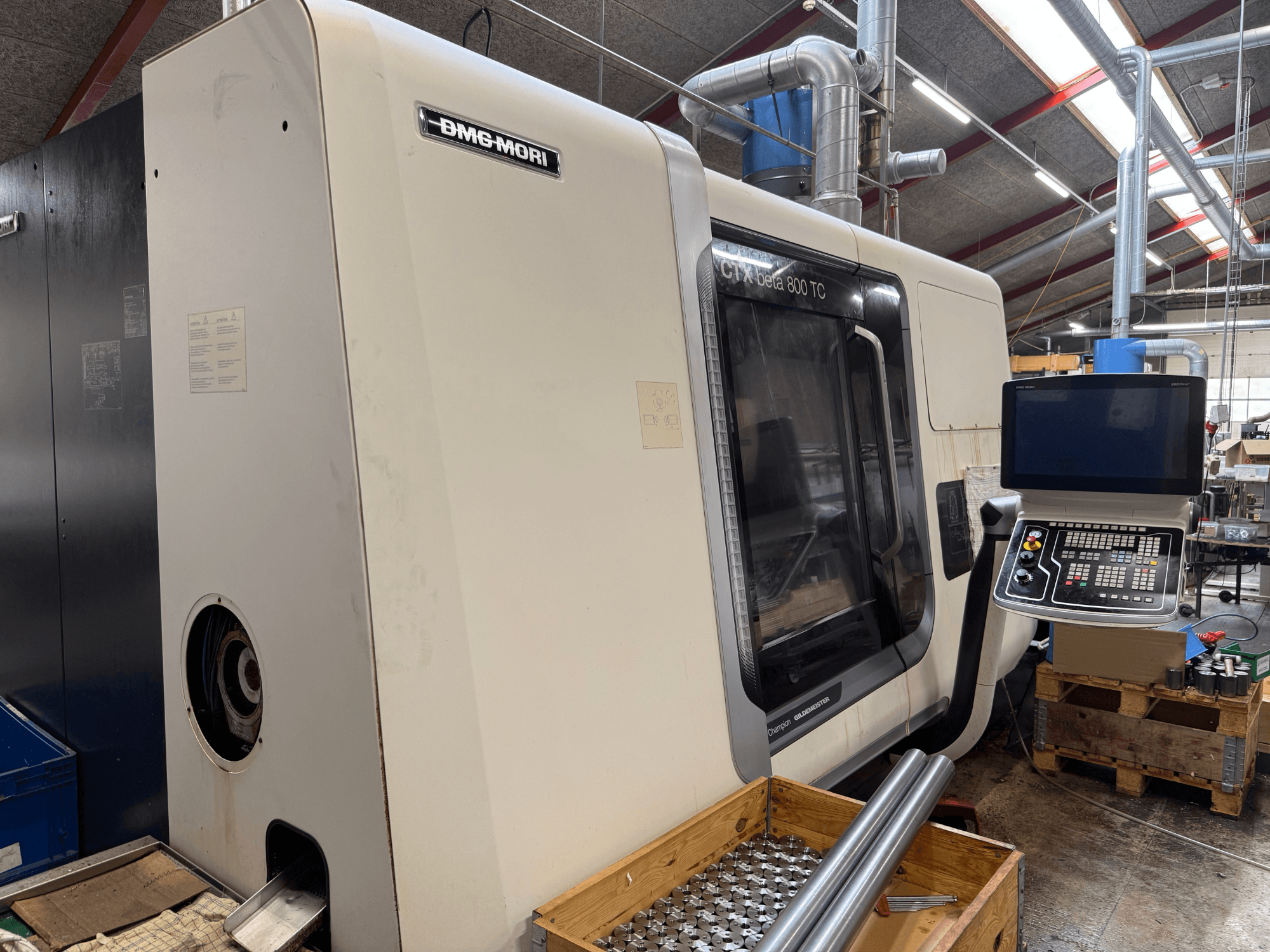 Máquina DMM-MORI CTX beta 800 TC, vista frontal, com painel de controlo, ferramentas e hastes de metal numa palete de madeira.