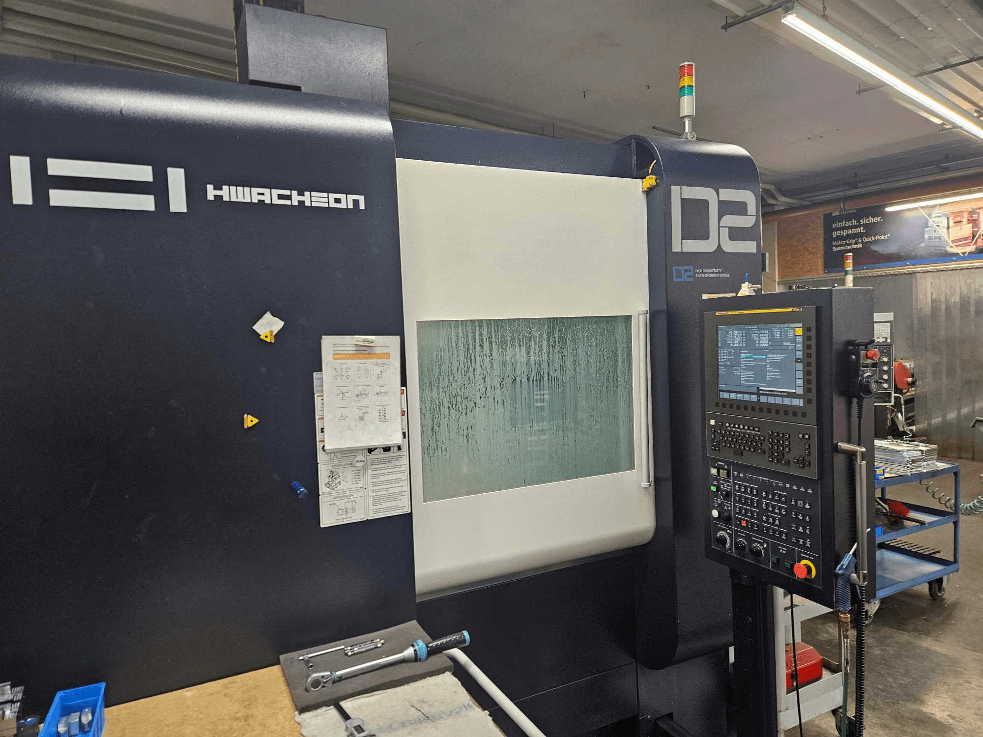 Centro de maquinagem vertical HWACHEON D2-5AX 12k, vista frontal que mostra o painel de controlo e a janela de visualização embaciada.