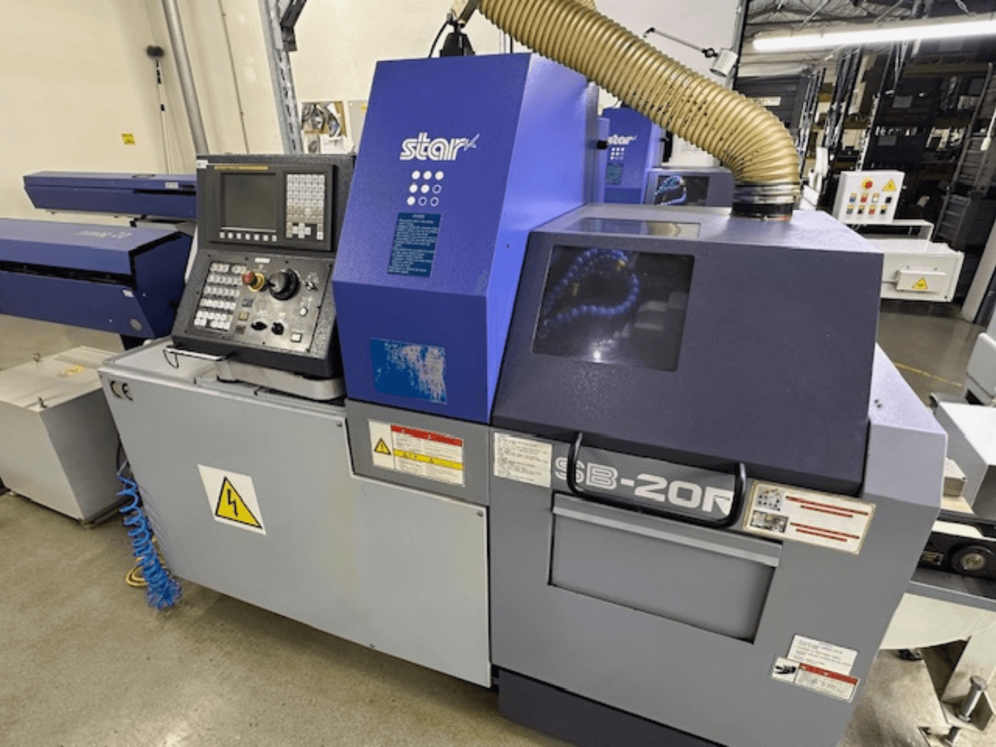 Máquina de tornear Star SB-20R tipo N de tipo suíço, vista frontal, com painel de controlo, mangueira de aspiração e símbolos de perigo visíveis.