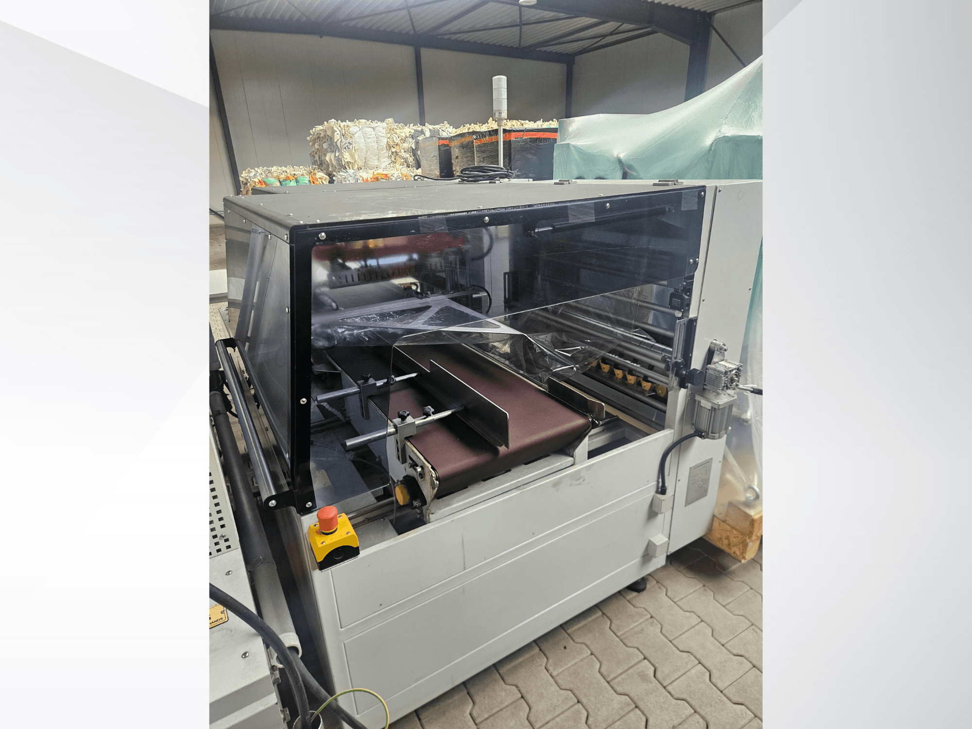Máquina AMTEC Speed Shrink 40 PRO, vista frontal, apresentando um mecanismo de processamento de plástico com uma cobertura de segurança transparente e uma correia transportadora.