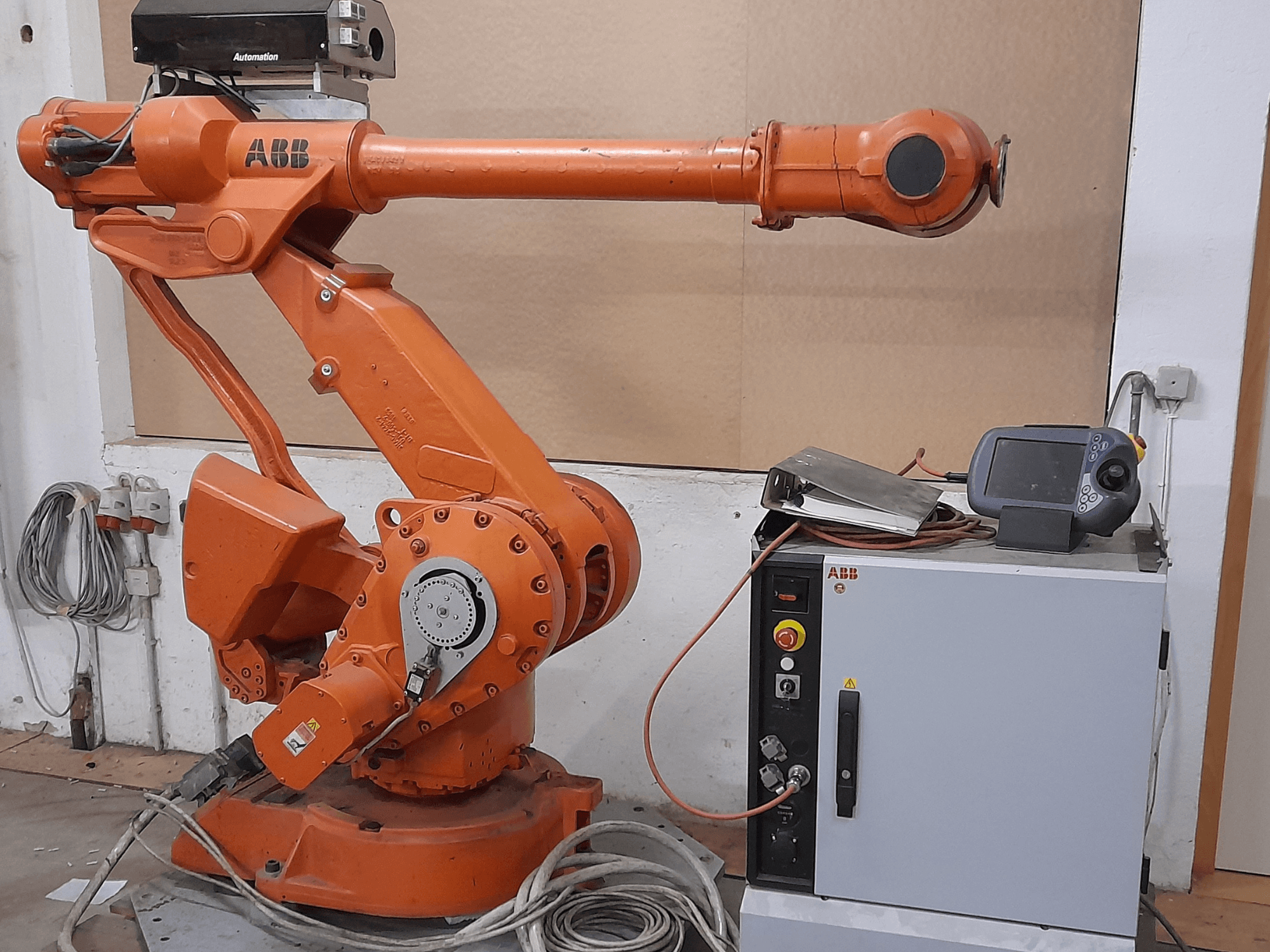 Braço robótico ABB laranja com painel de controlo, vista frontal, apresentando cabos e equipamento de automação.