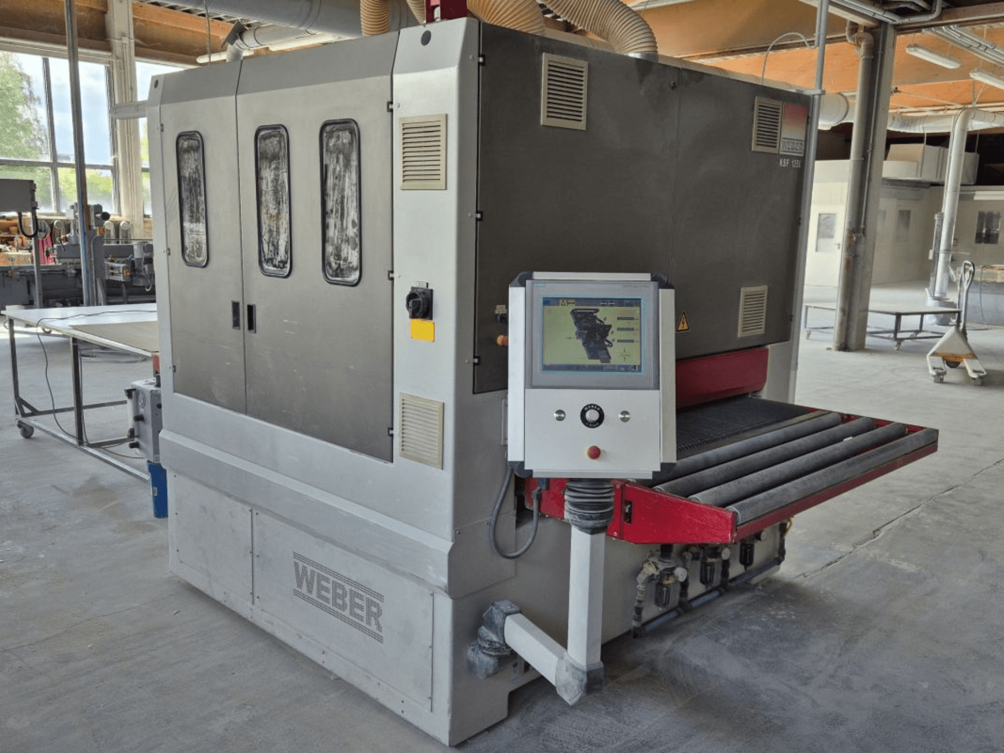 Lixadeira de cinta WEBER KSF-3-1350, vista lateral mostrando os controlos, o painel de alimentação e o tapete de lixagem. Ambiente industrial com máquinas visíveis.