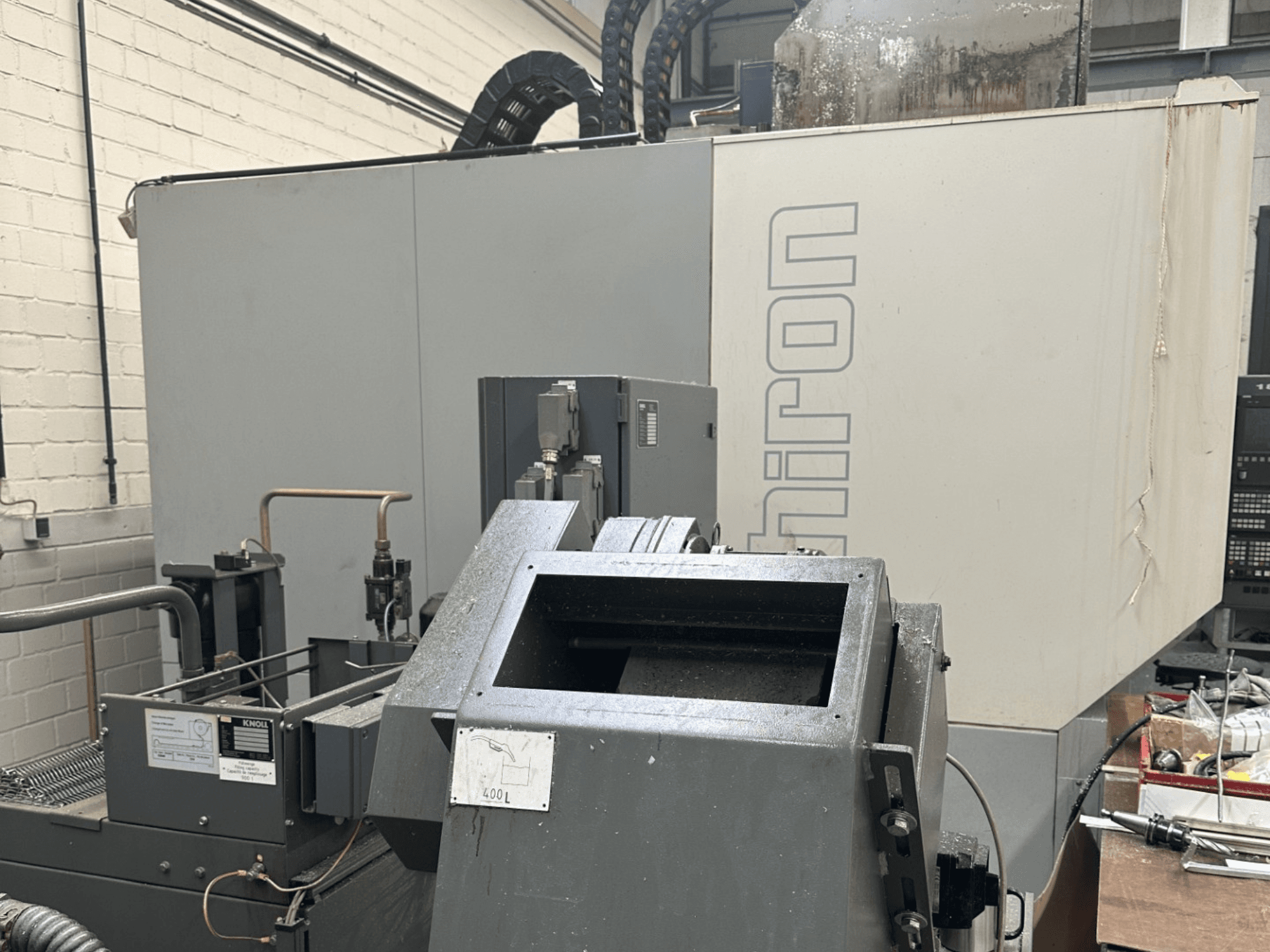 MARCA: Chiron; Vista frontal de uma máquina CNC industrial com exterior cinzento, painel de controlo visível e componentes da máquina em foco.