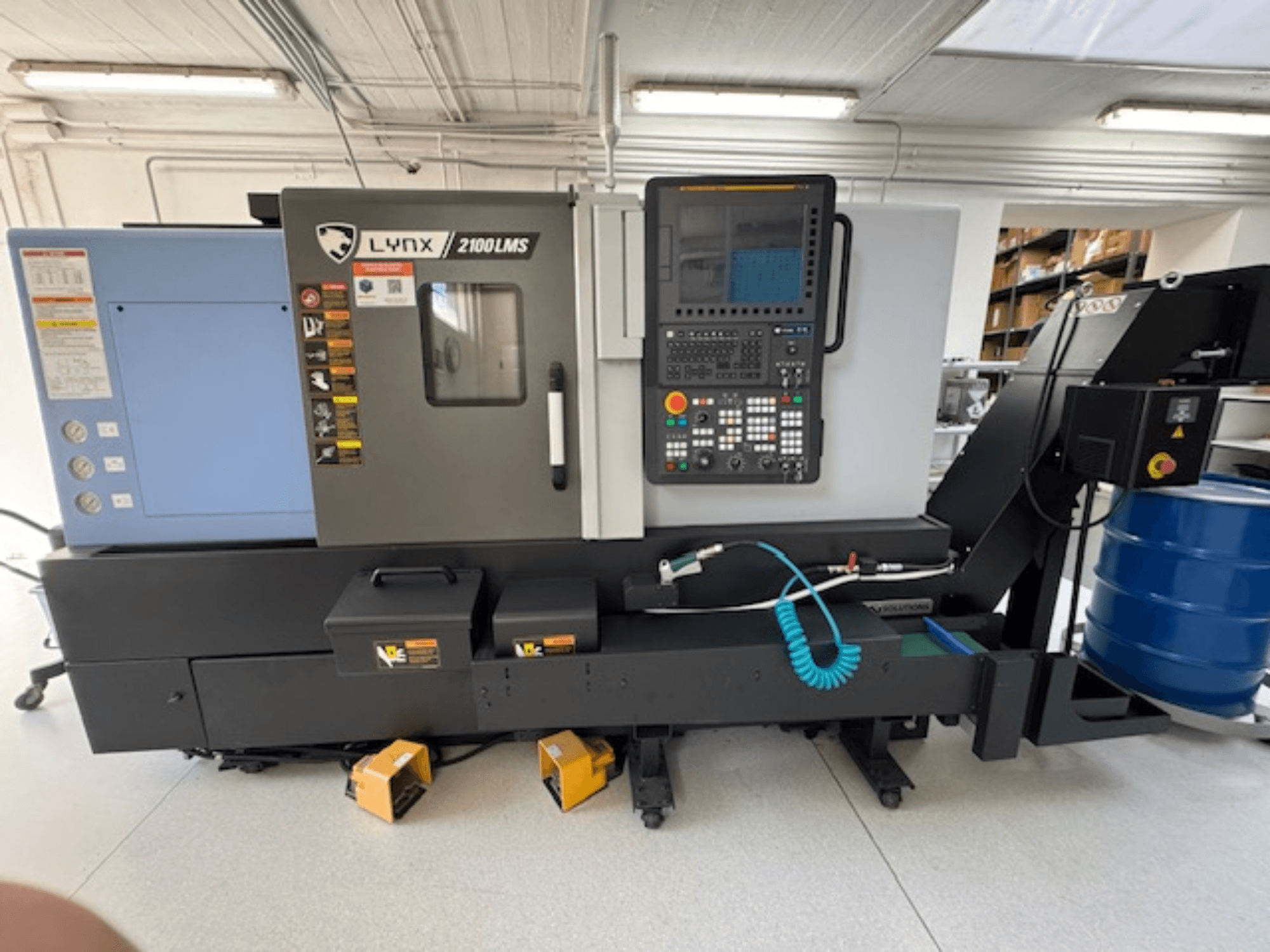 Torneiro horizontal DOOSAN LYNX 2100LMSA, vista frontal, com painel de controlo, mangueira de refrigeração e caixas de arrumação.