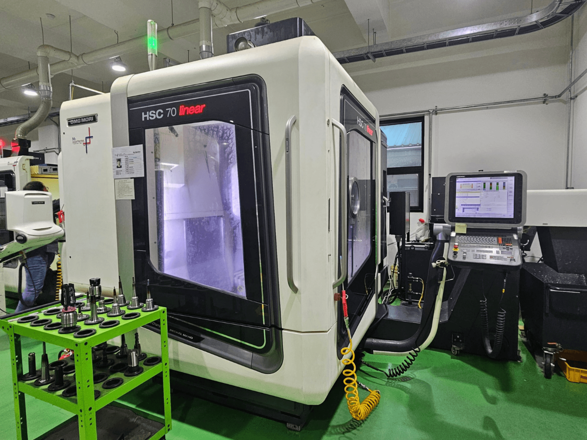 Centro de maquinação universal linear DMG MORI HSC 70, vista frontal, com suportes de ferramentas numa mesa verde e um painel de controlo visível.
