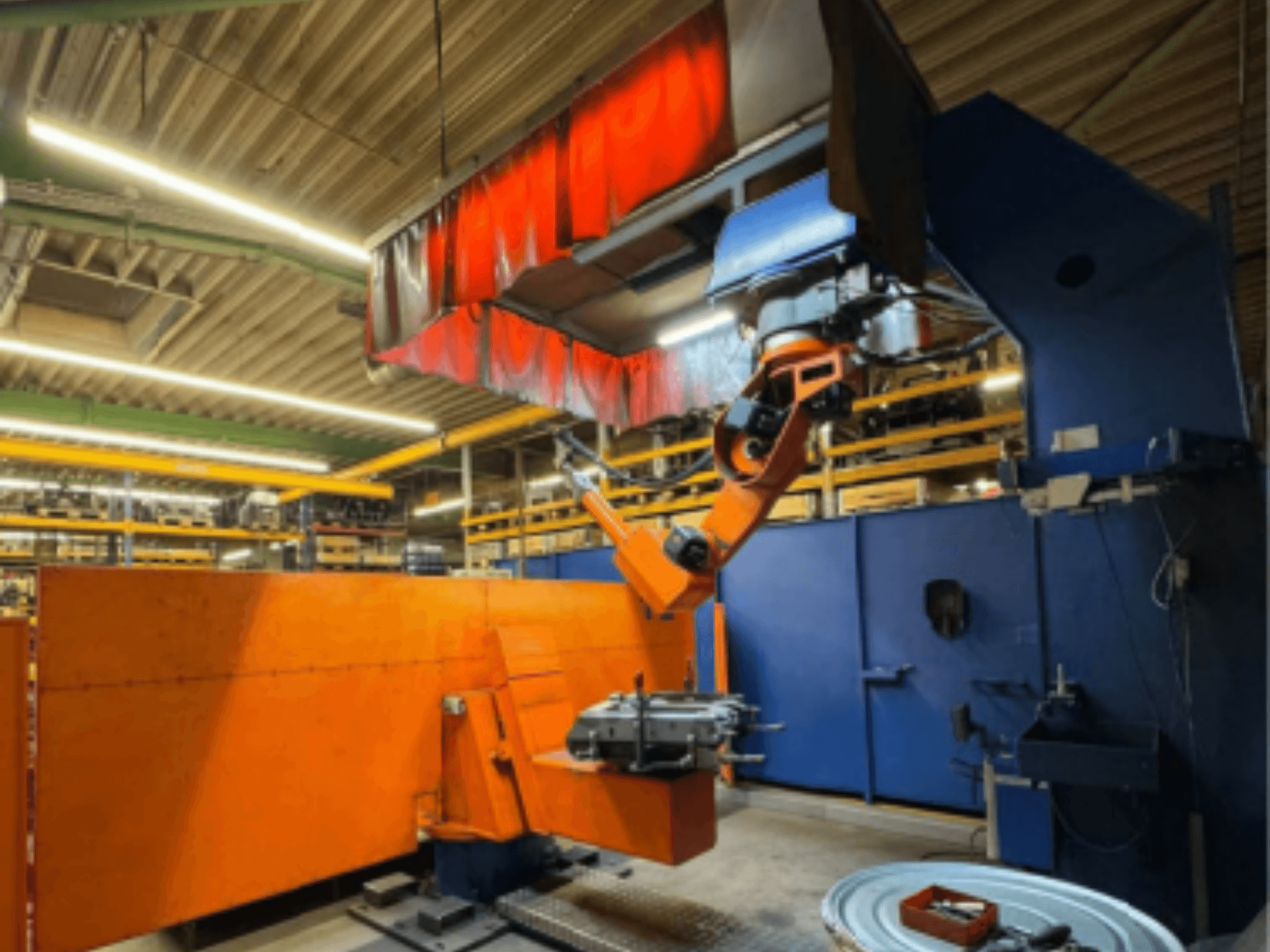 Robô de soldadura CLOOS Romat 350 e num ambiente industrial, vista frontal com braço laranja, segurando uma peça de trabalho sob uma cortina suspensa.