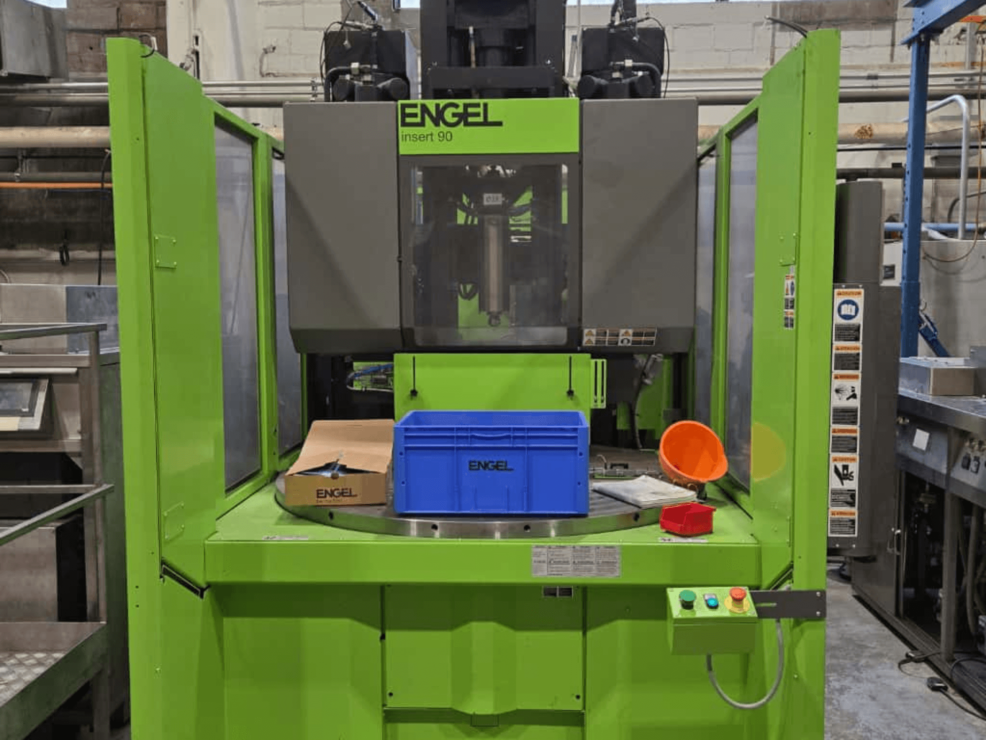 Vista frontal da máquina de moldagem por injeção ENGEL Insert 90 verde, com recipiente azul e painel de controlo com botões.