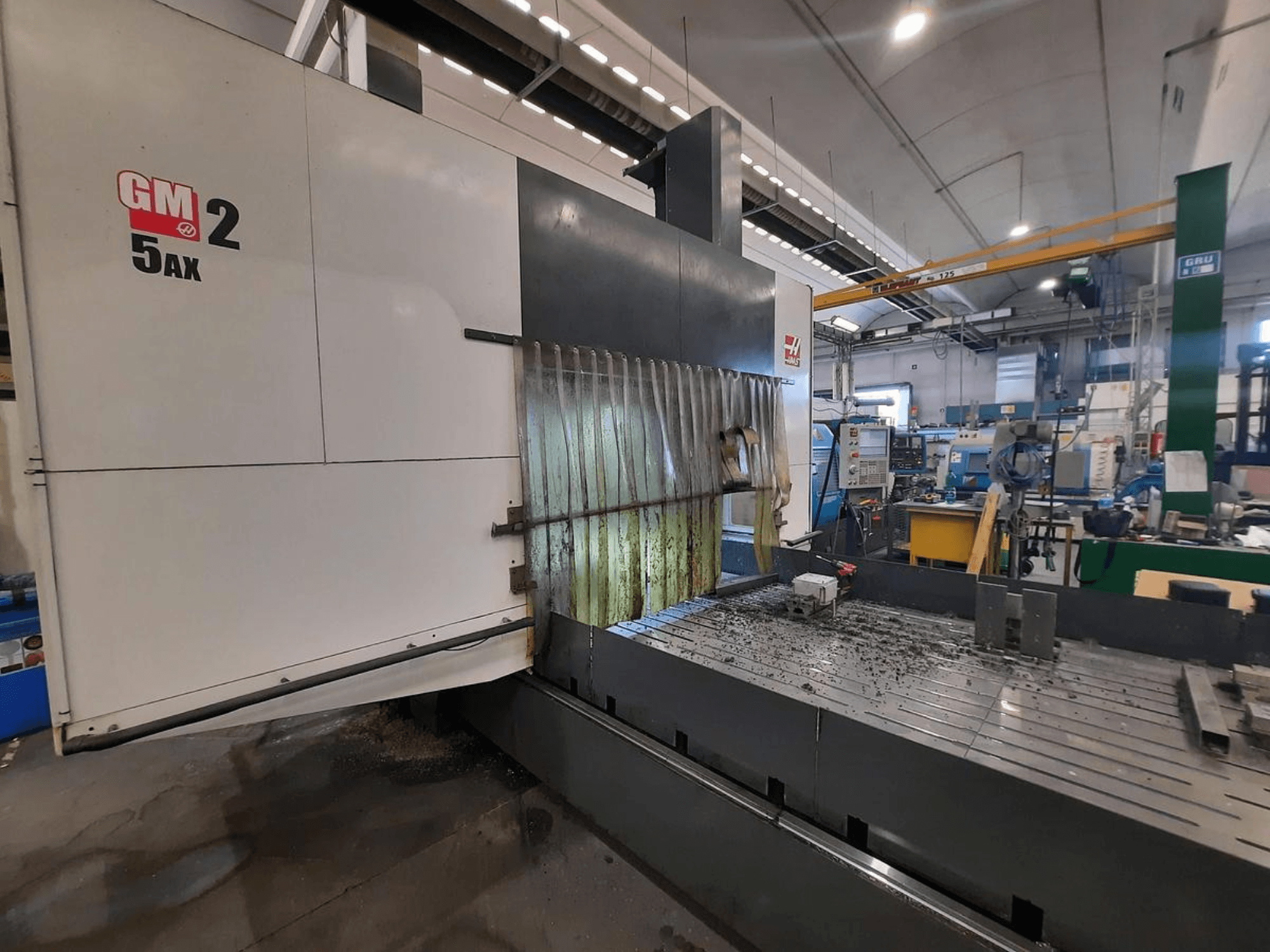 Máquina CNC GM 2 5AX numa oficina, vista lateral, mostrando a área operacional e aparas de metal na superfície da mesa.