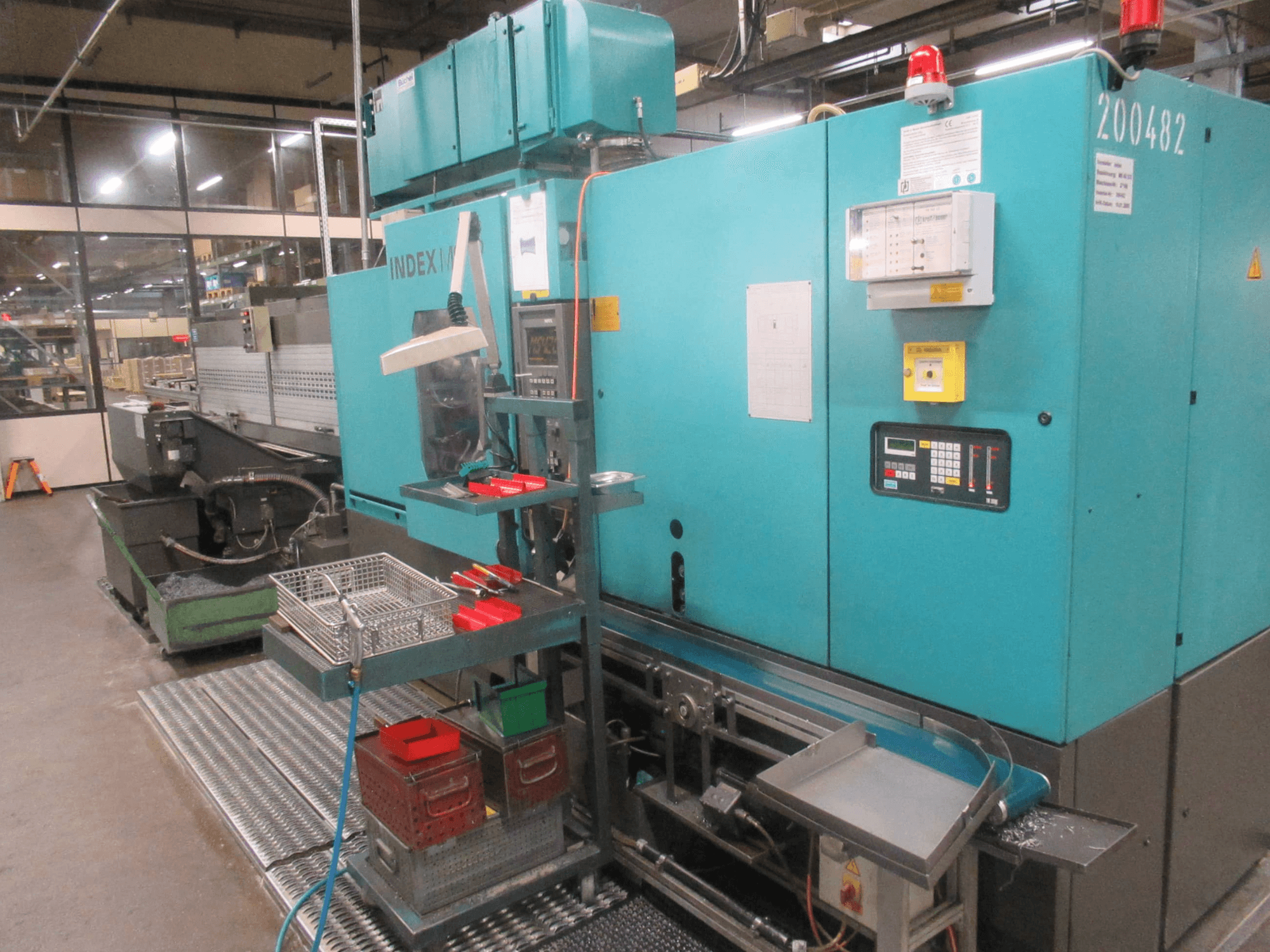 Máquina INDEX turquesa na fábrica, vista lateral; possui painel de controlo, armazenamento de ferramentas e transportador. Caixas, mangueiras e alarmes visíveis.