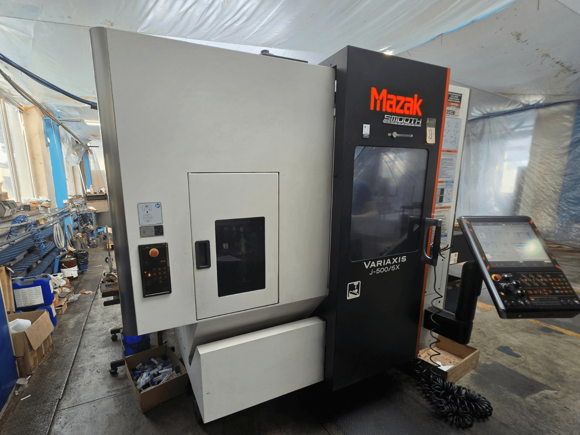 Centro de maquinagem vertical Mazak VARIAXIS J-500/5X, vista lateral que mostra os comandos e o ecrã, instalado num ambiente de oficina.