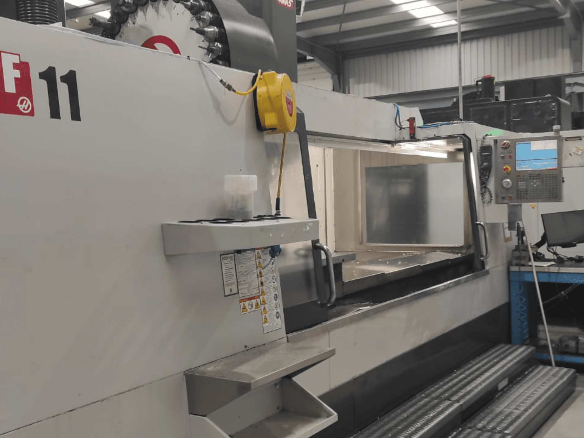 Maquinaria da Haas, modelo VF-11, mostrada de uma vista frontal com painel de controlo, grande área de trabalho e etiquetas de segurança visíveis.