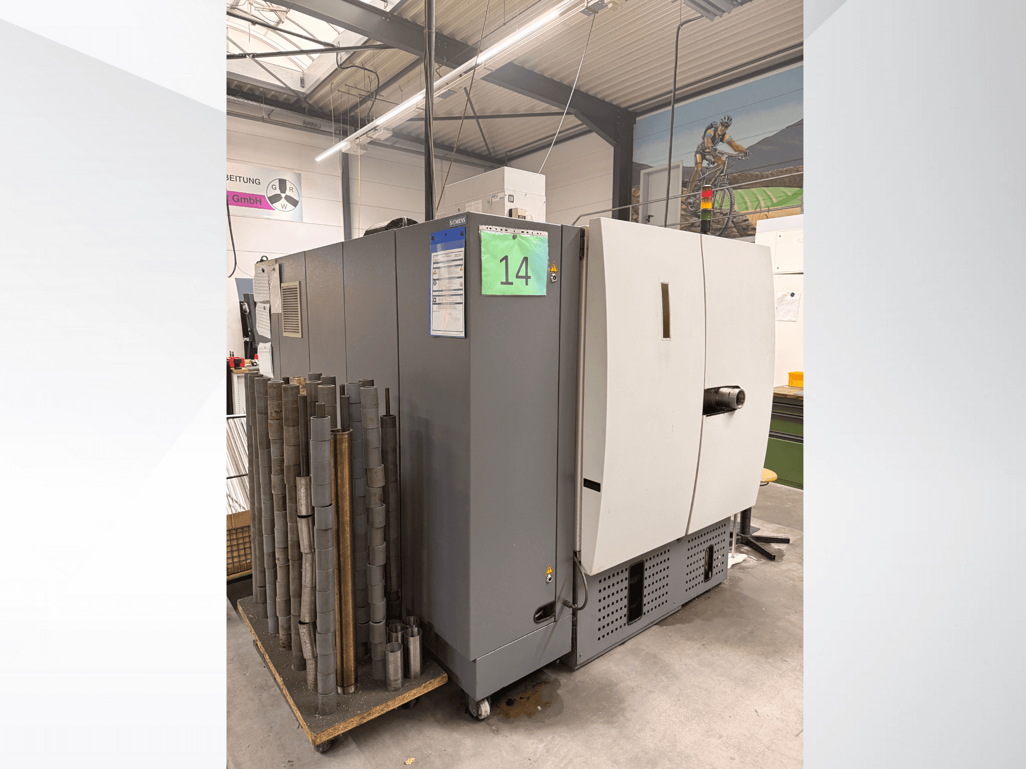 Máquina industrial cinzenta da Siemens, vista lateral, com stock de peças metálicas cilíndricas ao lado e etiquetas de equipamento visíveis.