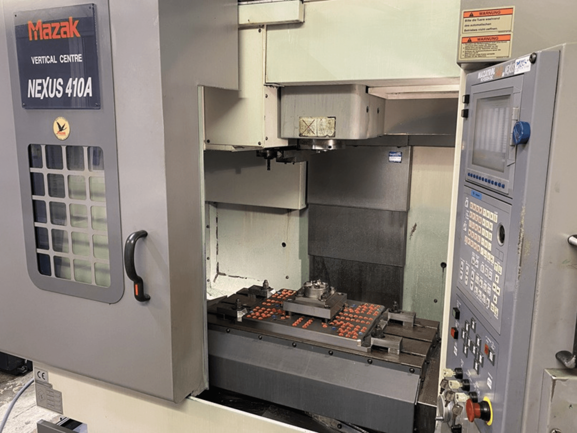 Centro de maquinação vertical Mazak VCN410, vista frontal mostrando o painel de controlo e o interior da máquina com a peça de trabalho montada.