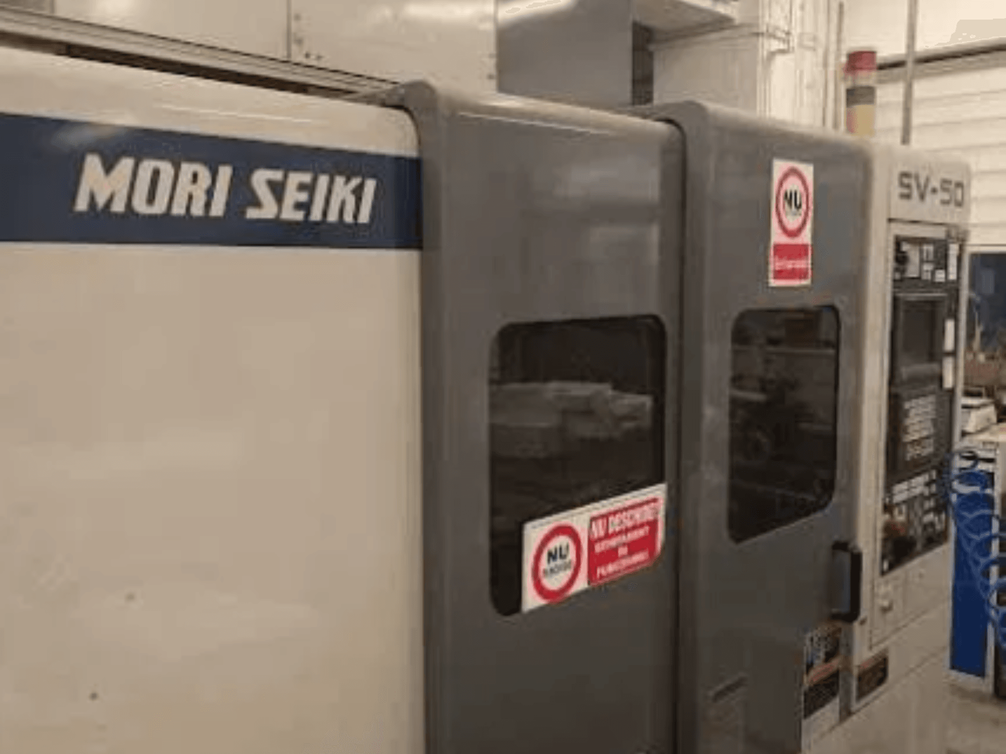 Máquina CNC MORI SEIKI SV-50, vista lateral; possui painel de controlo e autocolantes de aviso de segurança.