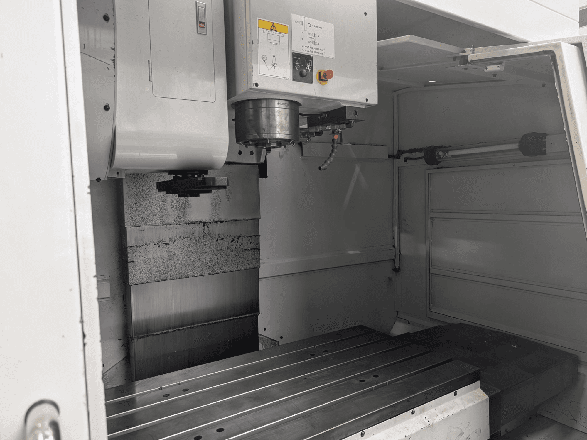 Vista interior de uma fresadora CNC com uma grande mesa de trabalho em metal e um motor de fuso, mostrando componentes pormenorizados e o painel de controlo.