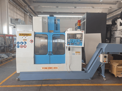Centro de maquinagem vertical SUPERMAX YCM-VMC-85A numa fábrica, com painel de controlo e autocolantes de segurança, vista frontal.