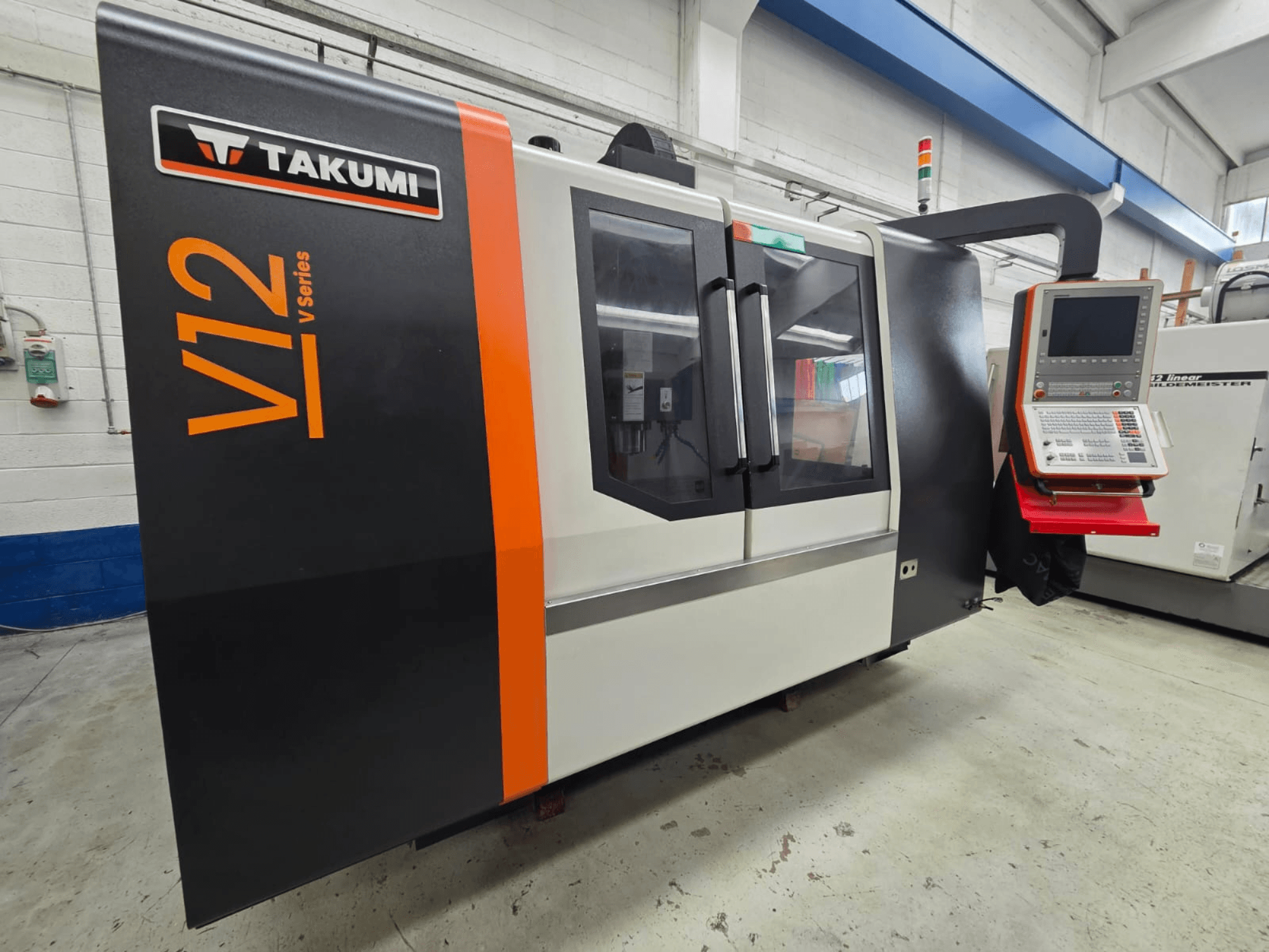 Vista frontal da fresadora CNC Takumi V12 da série V, com painel de controlo e grandes janelas de vidro.