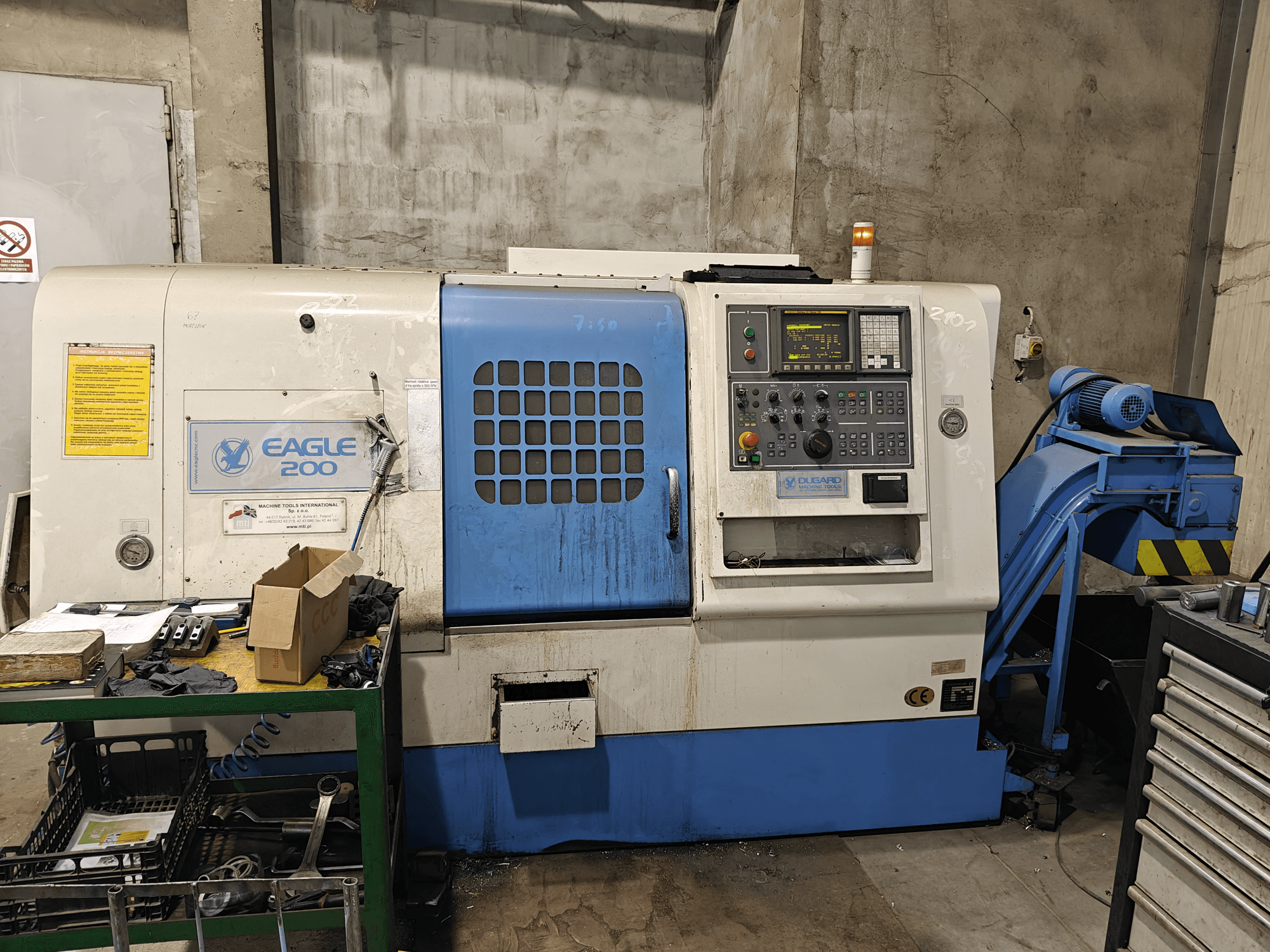 Máquina CNC Eagle 200, vista frontal com painel de controlo, várias ferramentas num carrinho verde. Desgaste visível e luzes operacionais.