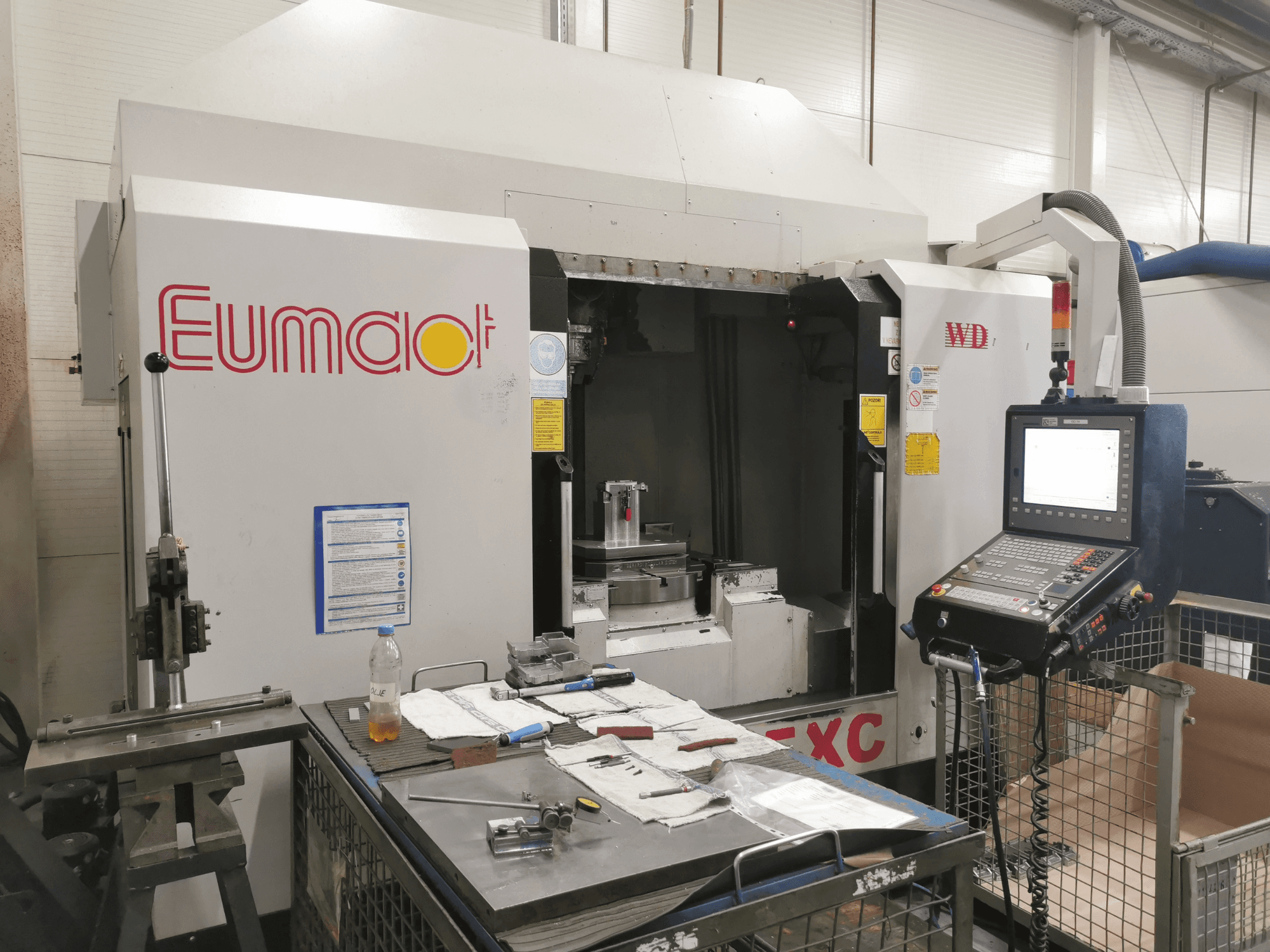 Vista frontal de uma máquina CNC Eumdaot com painel de controlo, ferramentas e uma superfície de trabalho repleta de componentes e materiais.