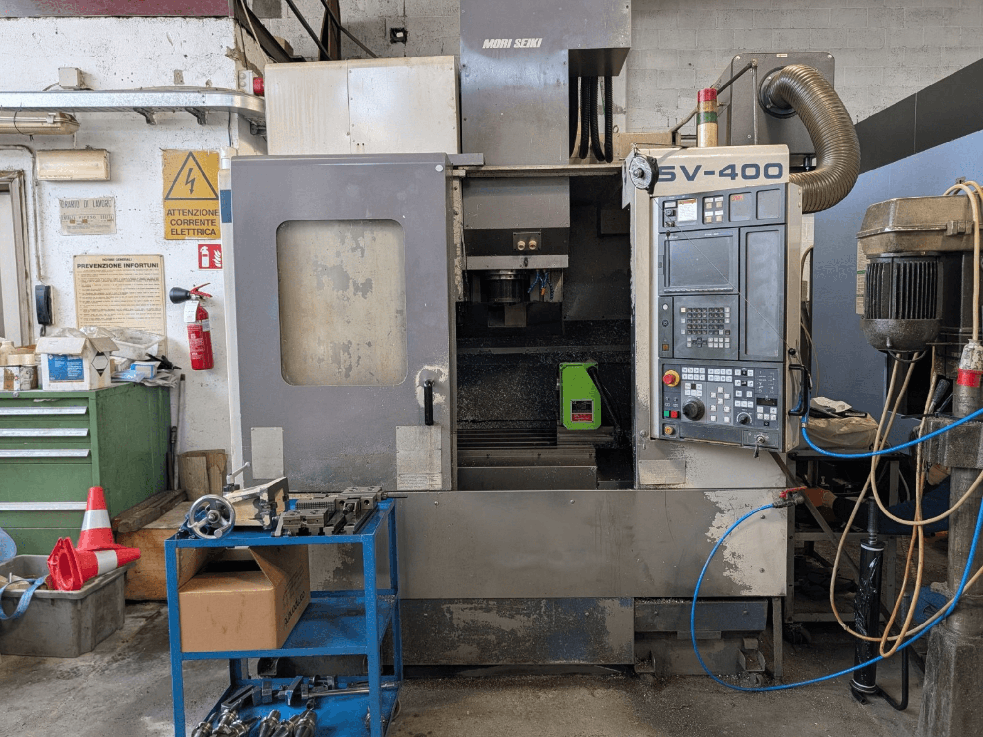 Centro de maquinação vertical MORI SEIKI SV-400 numa oficina, vista lateral, mostrando o painel de controlo e as máquinas-ferramentas próximas.