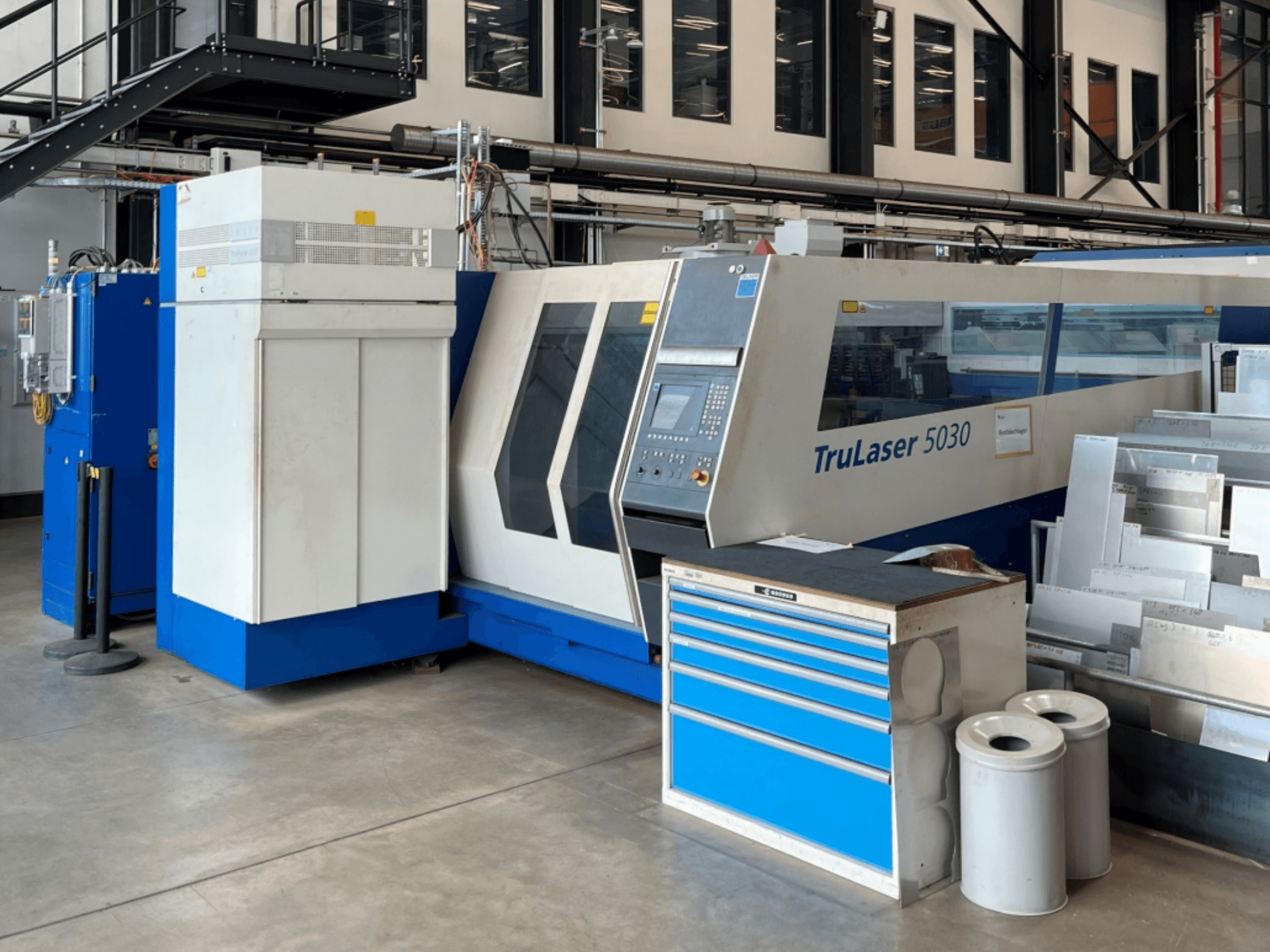 Máquina de corte a laser de CO₂ TRUMPF TruLaser 5030, vista de frente, com o painel de controlo e a bancada de trabalho nas proximidades, com chapas metálicas.