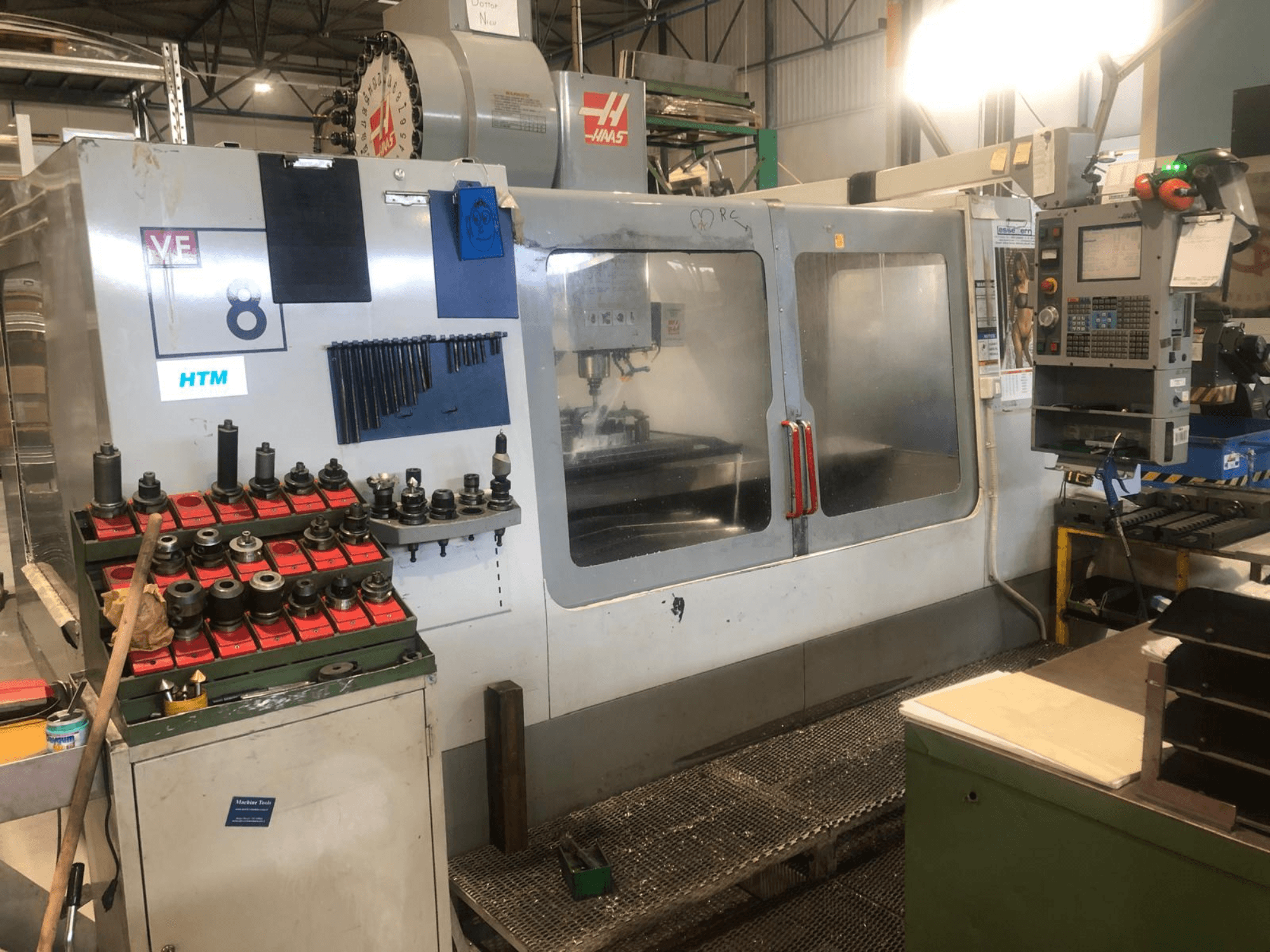 Máquina CNC Haas numa oficina, vista frontal; apresenta ferramentas e controlos, com detalhes visíveis do modelo e suportes de ferramentas organizados.