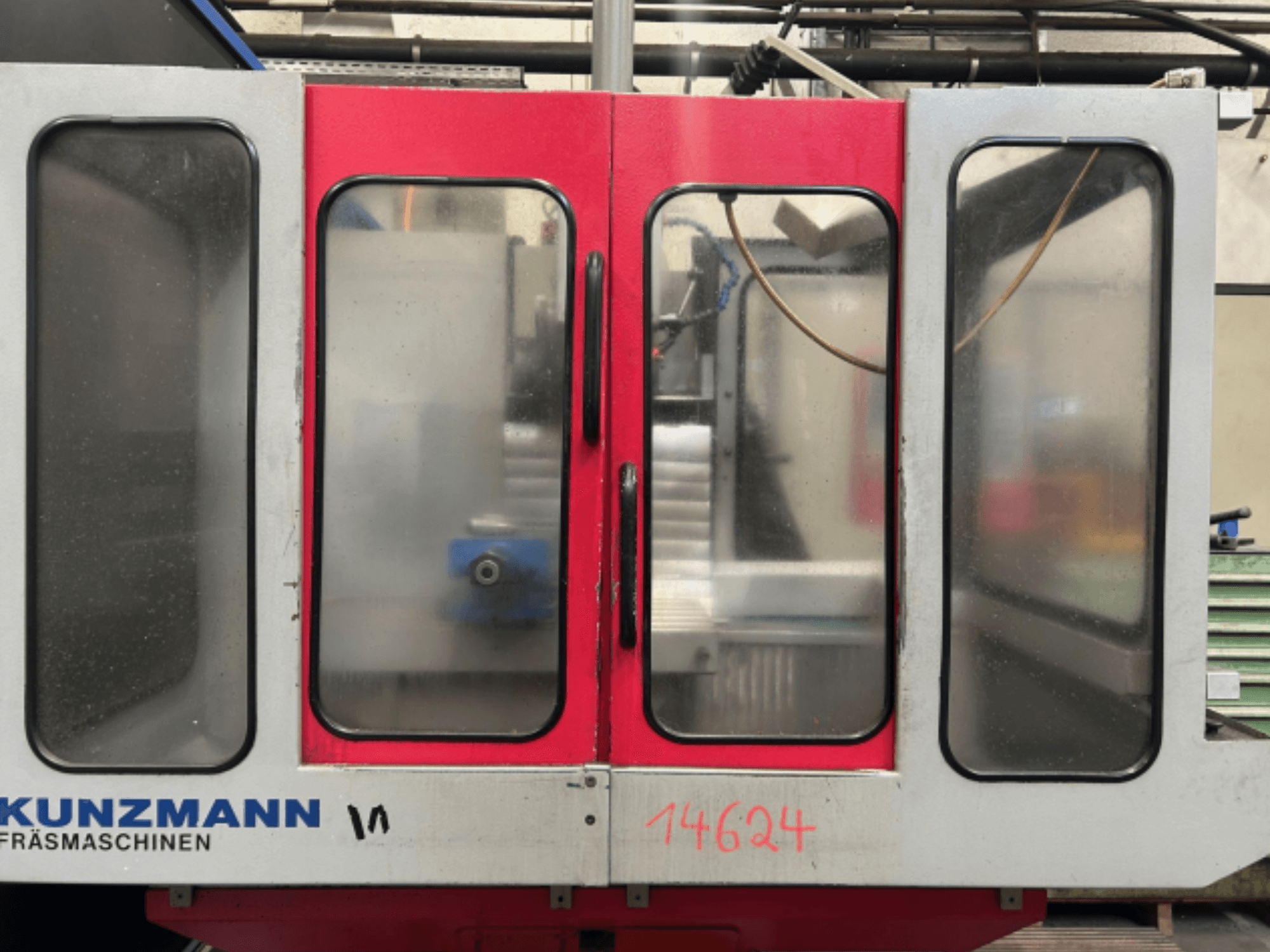 Vista frontal de uma máquina CNC KUNZMANN, com painéis vermelhos e cinzentos, com um painel de controlo visível e um suporte de ferramentas no interior.