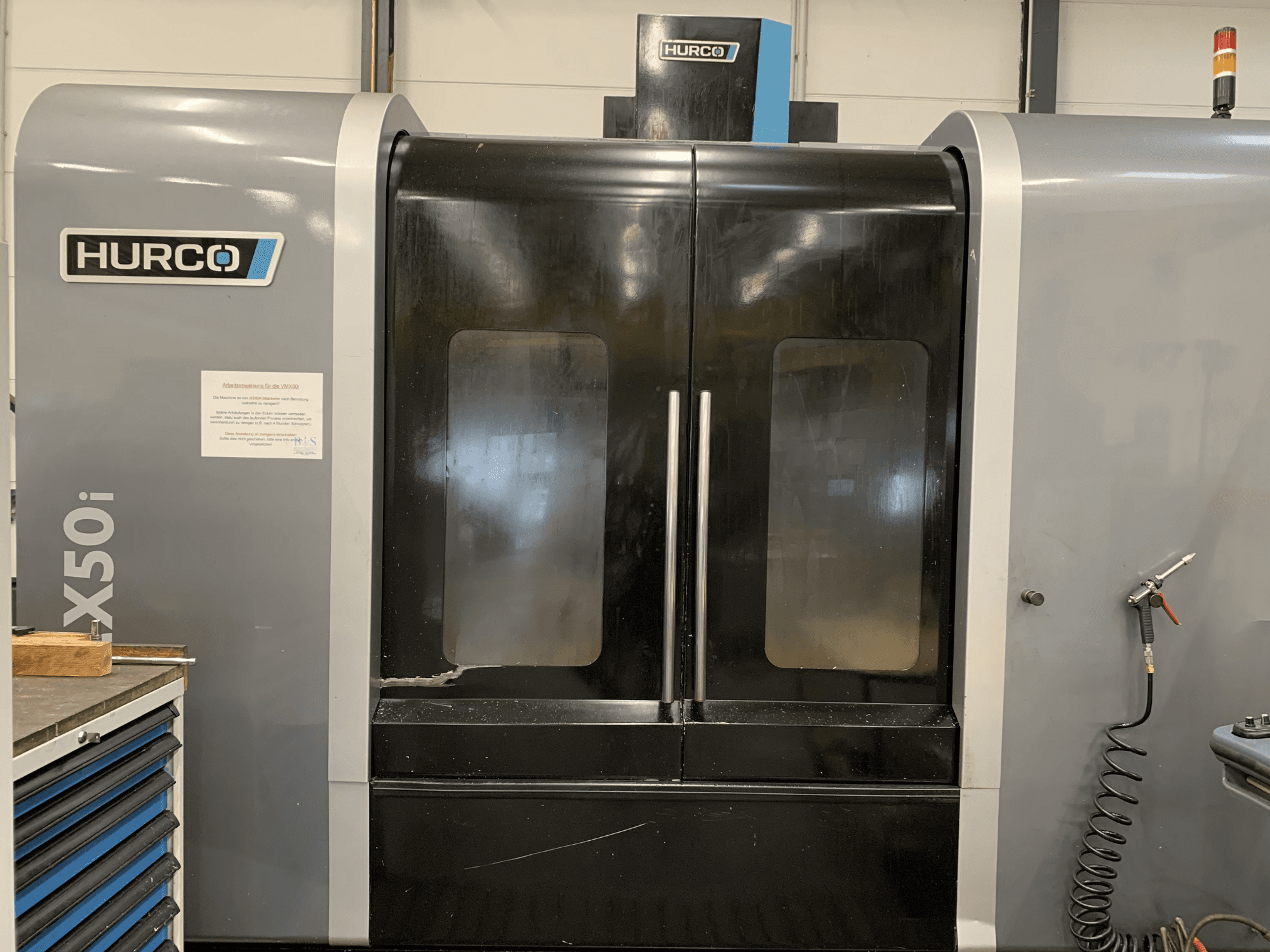 Vista frontal de uma máquina CNC Hurco VX50i com duas portas, mostrando os controlos técnicos e um sinal de manutenção na lateral.