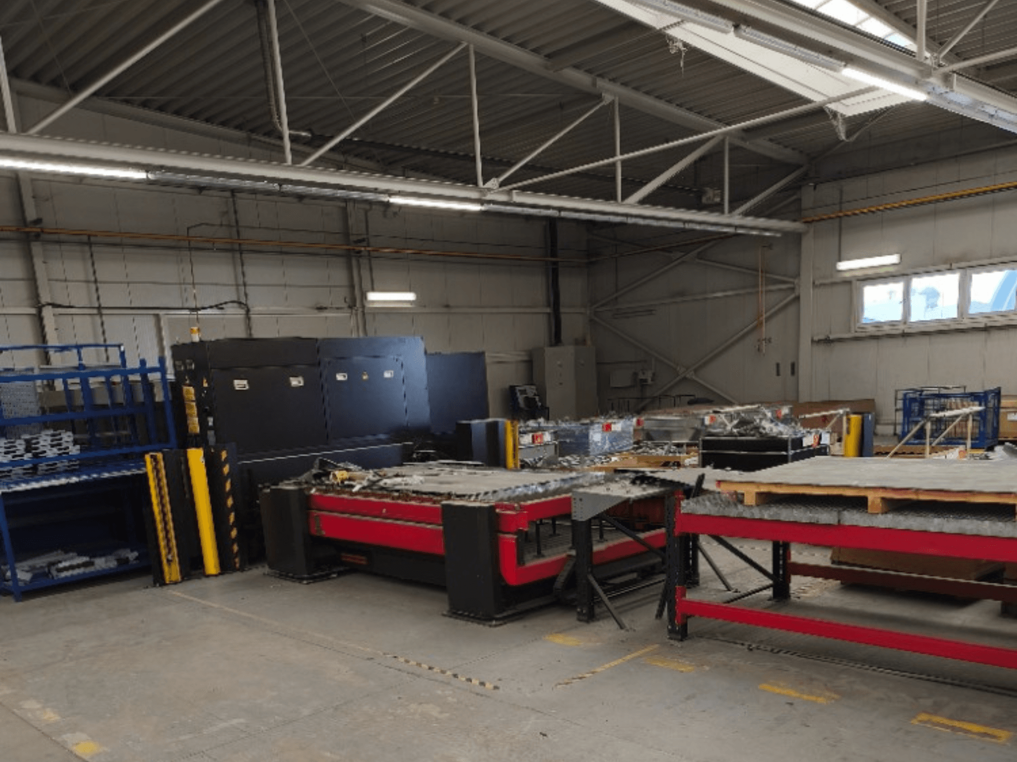 Máquina de corte a laser de fibra AMADA LCG 3015 AJ numa oficina, vista de frente, com prateleiras de armazenamento e materiais ao fundo.