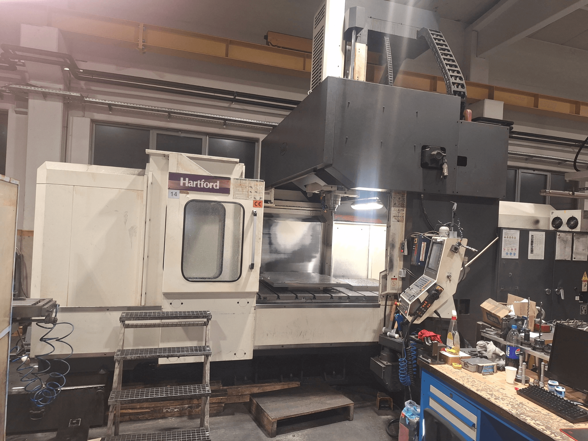 Máquina CNC Hartford, vista frontal, com controlos técnicos, degraus de acesso e uma área de mesa de trabalho, numa oficina.