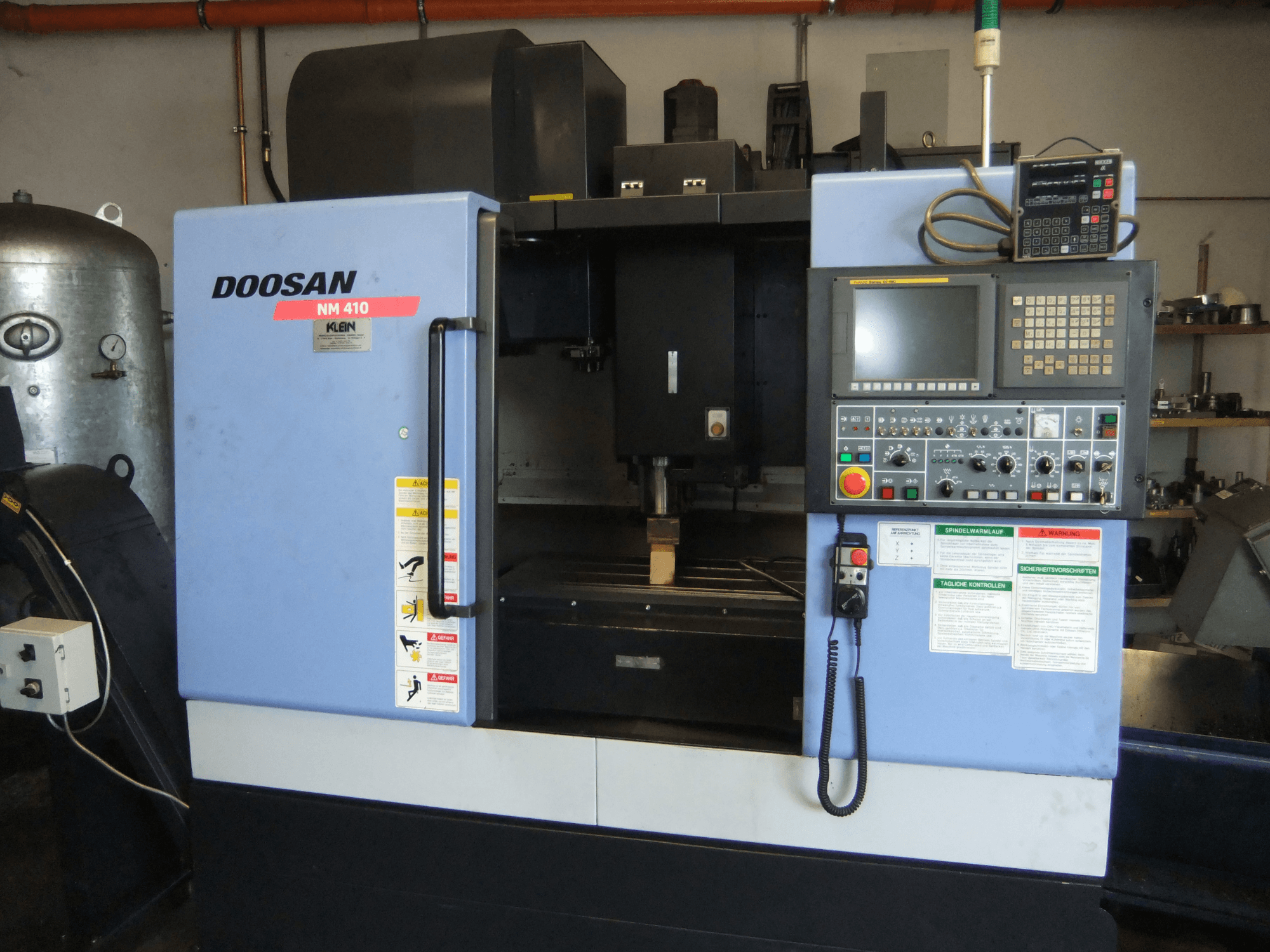 Centro de maquinagem vertical DOOSAN NM 410, vista frontal, mostrando o painel de controlo, a área do fuso e as etiquetas de segurança.