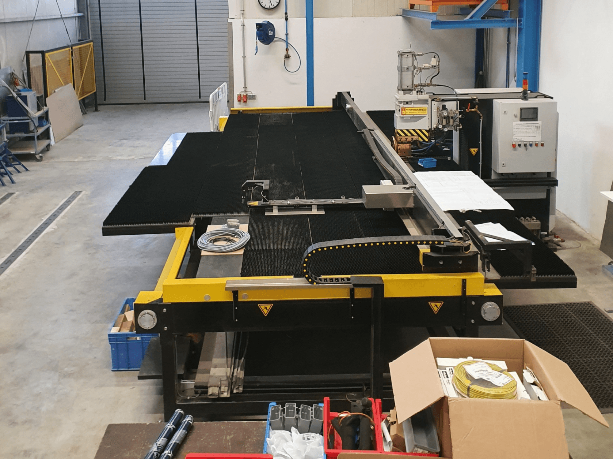 Máquina de corte industrial com caraterísticas amarelas e pretas, vista frontal, mostrando controlos e acessórios num ambiente de fábrica.