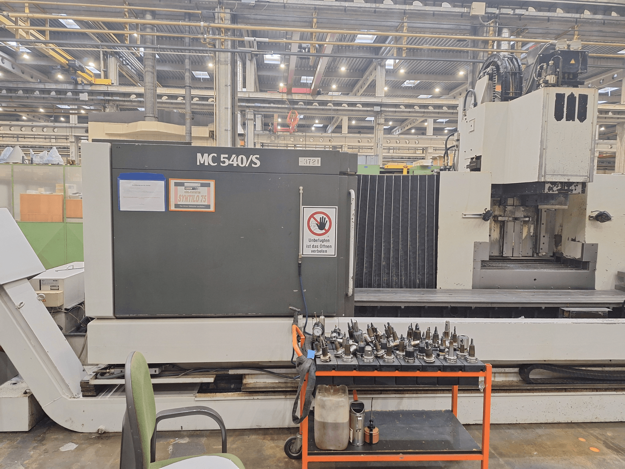 Centro de maquinação vertical STAMA MC 540S numa oficina, vista lateral com suportes de ferramentas num carrinho próximo.