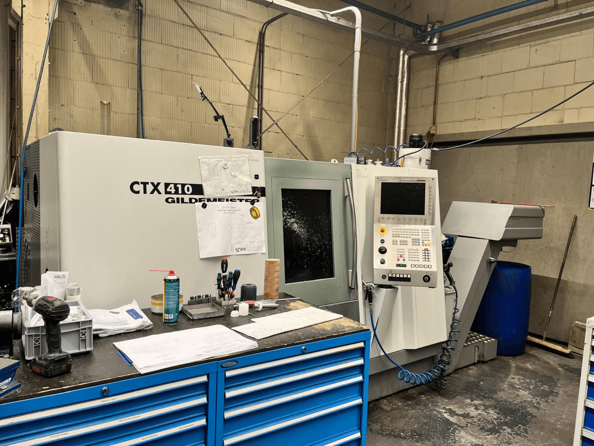 Máquina CNC Gildemeister CTX 410 numa oficina, vista frontal, com um painel de controlo e várias ferramentas numa bancada de trabalho.