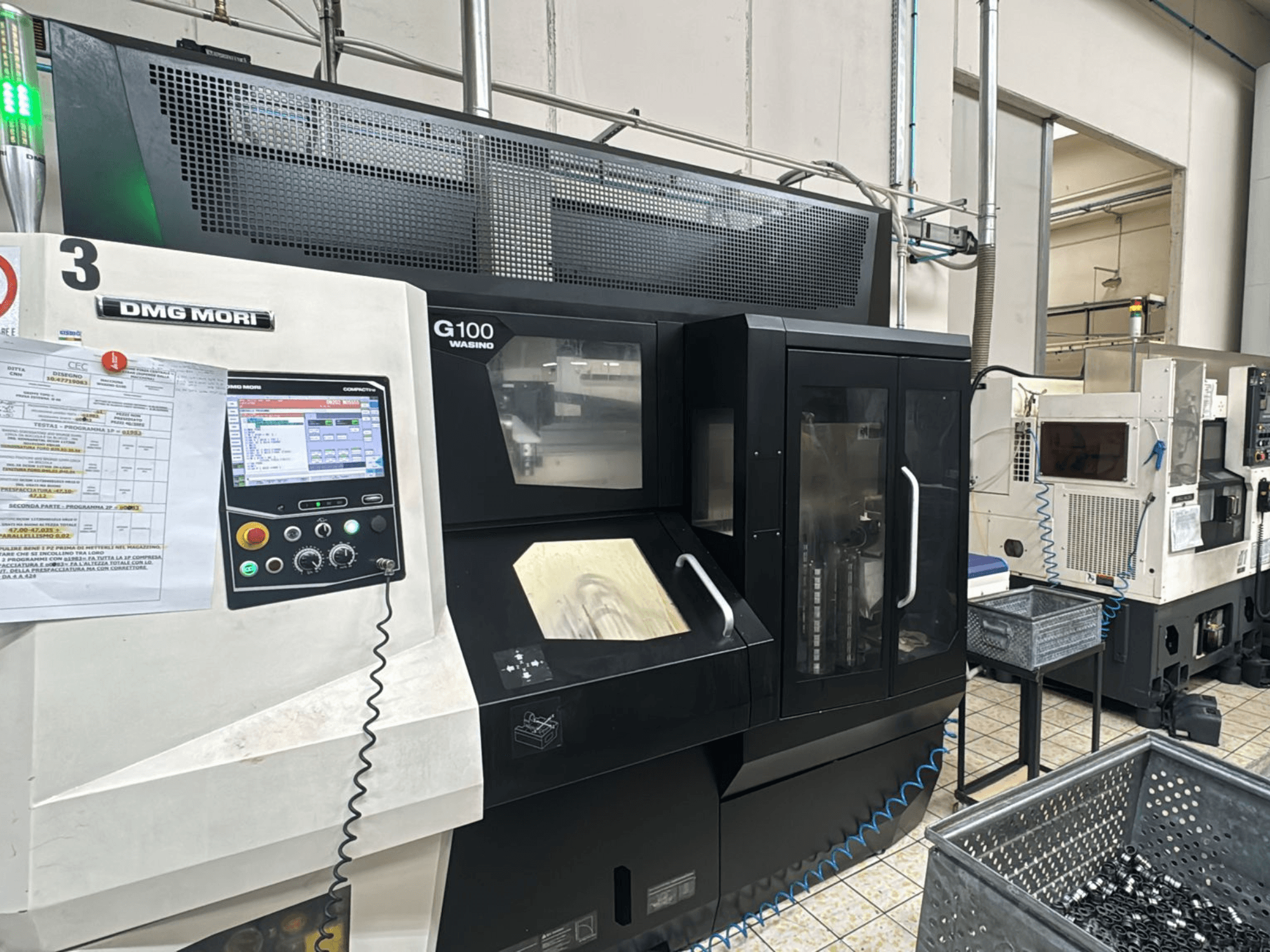 Máquina DMG MORI G100 numa fábrica, vista frontal, com um painel de controlo digital e caixas de armazenamento de ferramentas próximas.