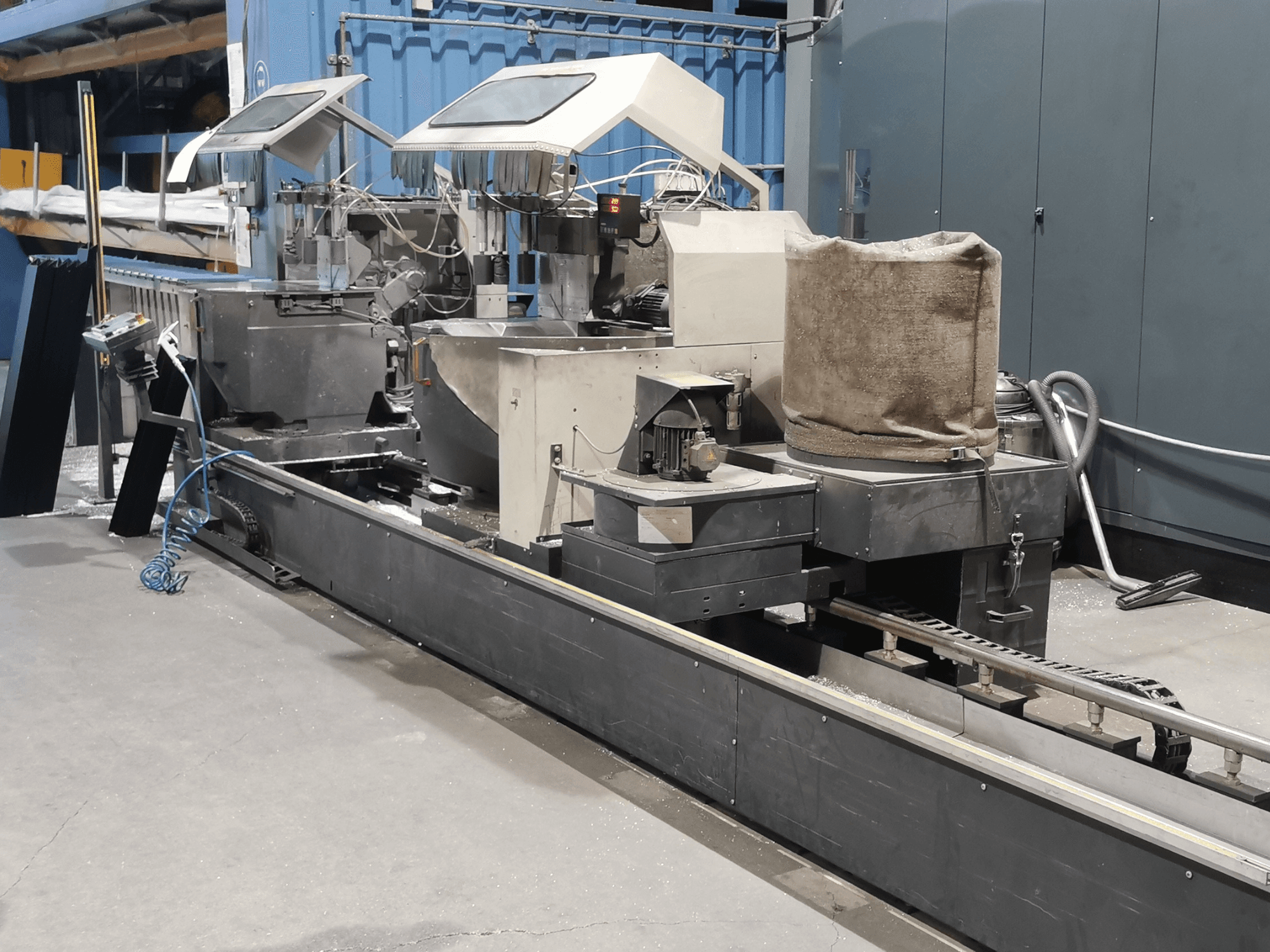 Máquina de processamento industrial vista de lado, com tremonhas, correia transportadora e painel de controlo, num ambiente de fábrica.