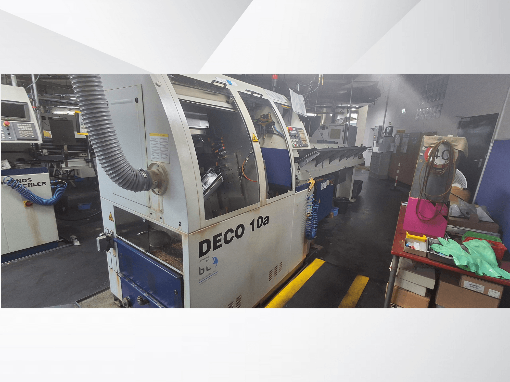 Máquina CNC DECO 10a numa oficina, vista frontal; possui um painel de controlo e um sistema de extração de poeiras, com uma bancada de trabalho próxima.