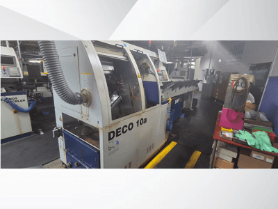 Máquina CNC DECO 10a numa oficina, vista frontal; possui um painel de controlo e um sistema de extração de poeiras, com uma bancada de trabalho próxima.