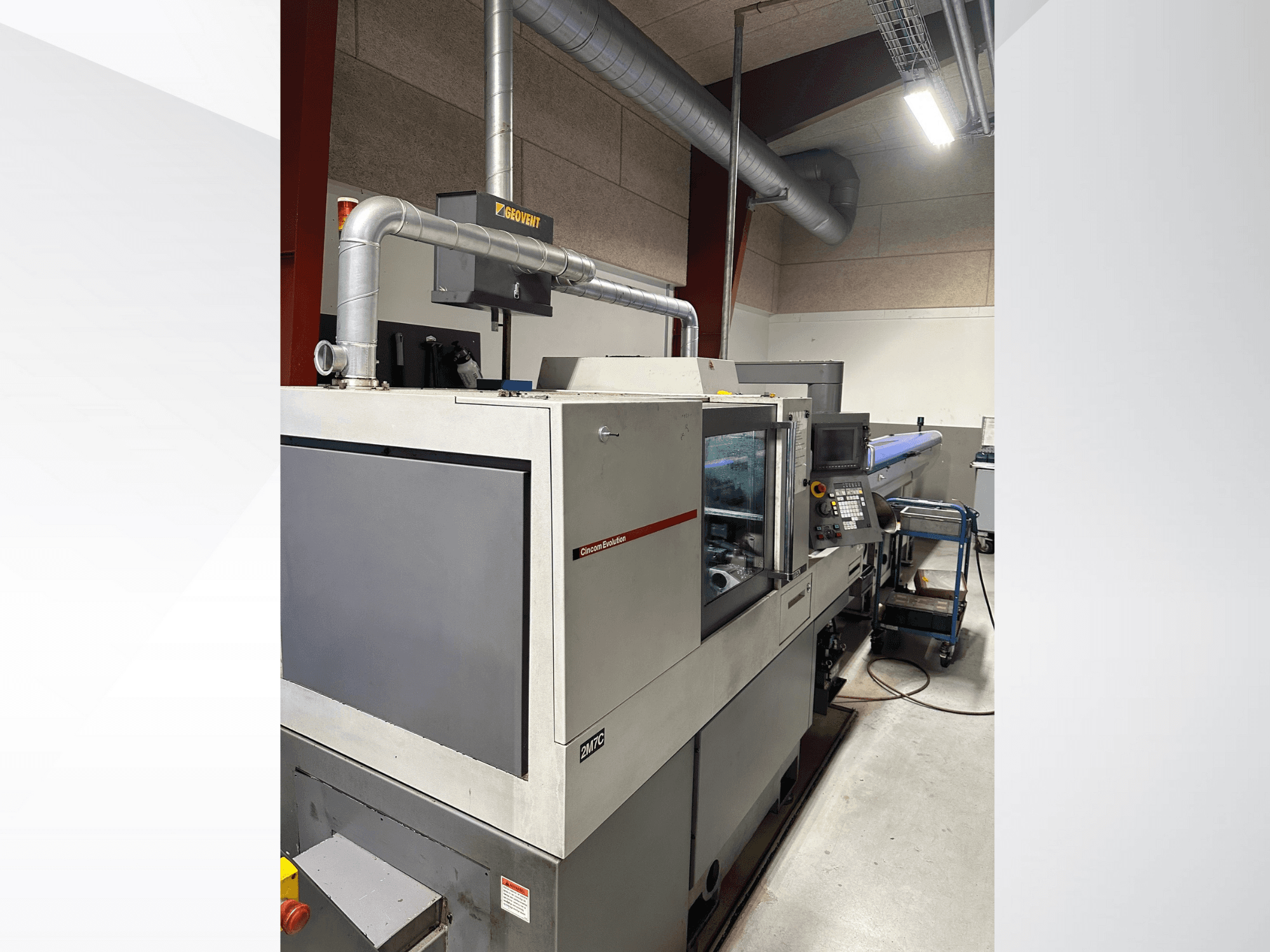 Máquina industrial da marca MVC, vista lateral com painel de controlo e condutas visíveis, inserida num ambiente de fábrica.