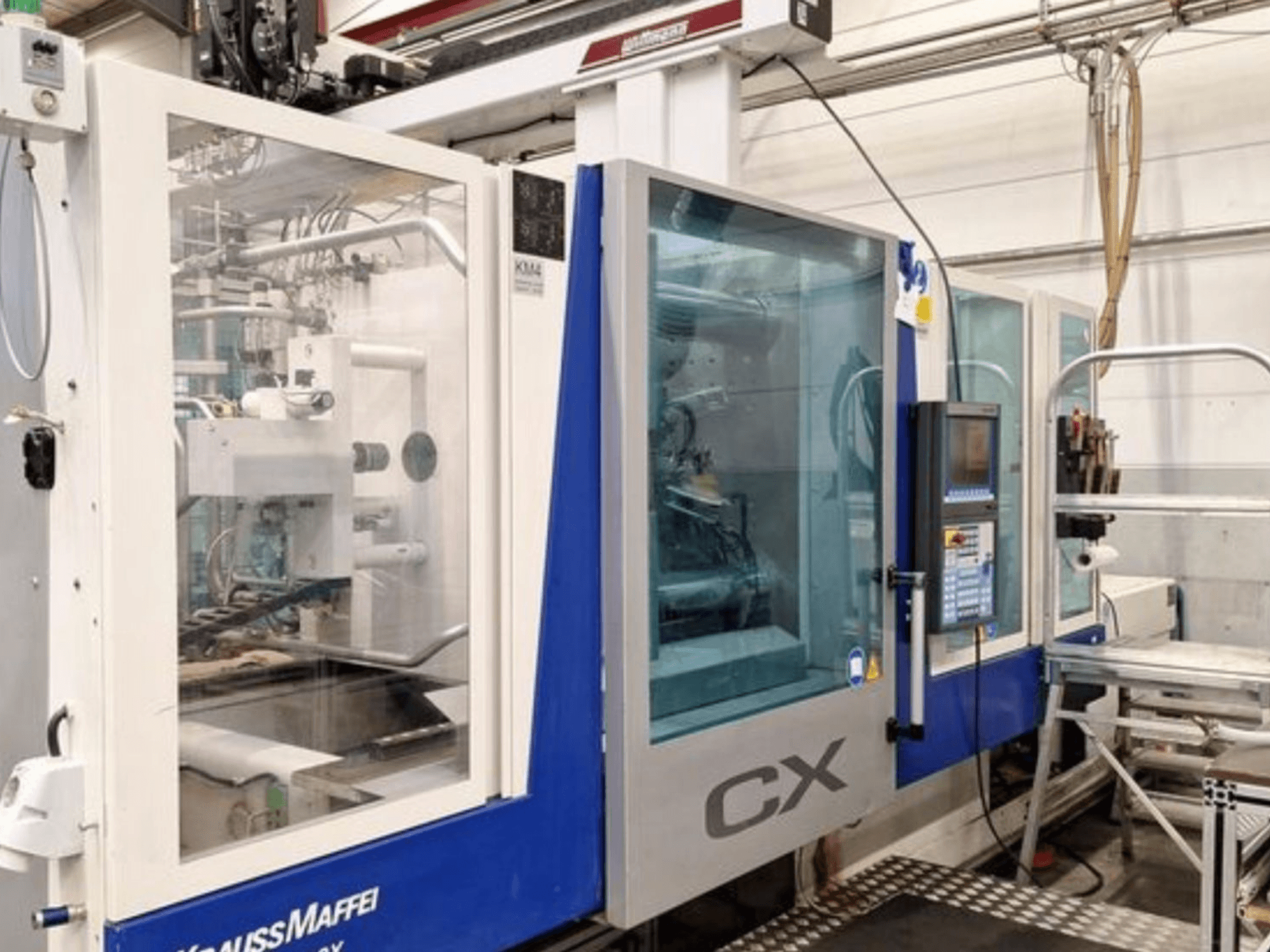 Máquina de moldagem por injeção hidráulica Krauss Maffei 300-1400 CX, vista lateral, mostrando o painel de controlo e a estrutura transparente.