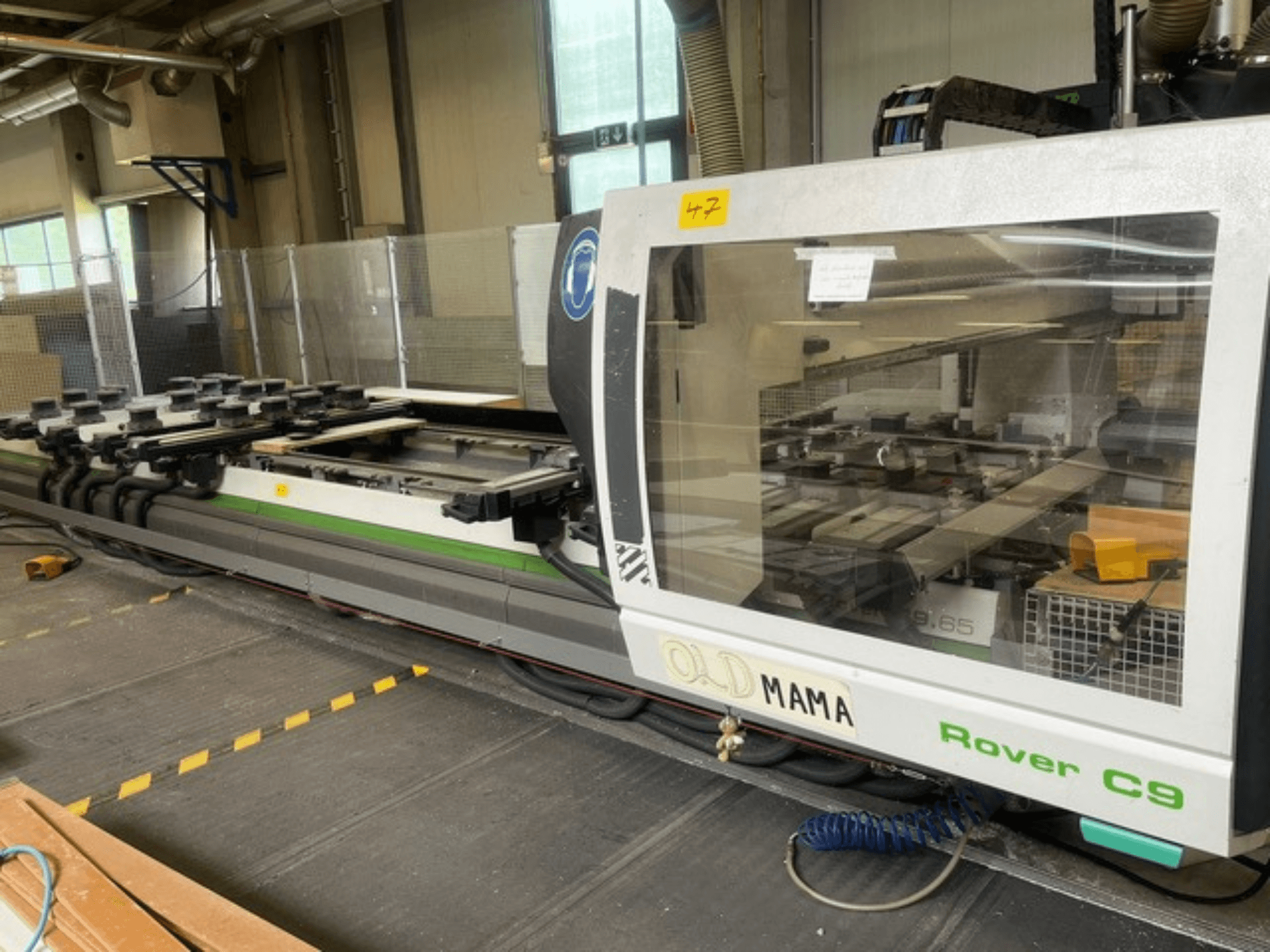 Tupia CNC BIESSE Rover C 9.65 numa oficina, vista lateral mostrando o painel de controlo e a mesa de trabalho com pinças.