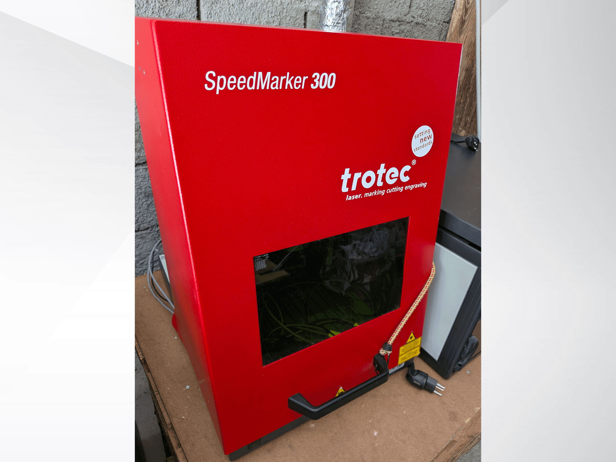 Máquina de marcação a laser SpeedMarker 300 vermelha da Trotec em vista frontal, com uma grande janela de visualização e ligação à corrente.
