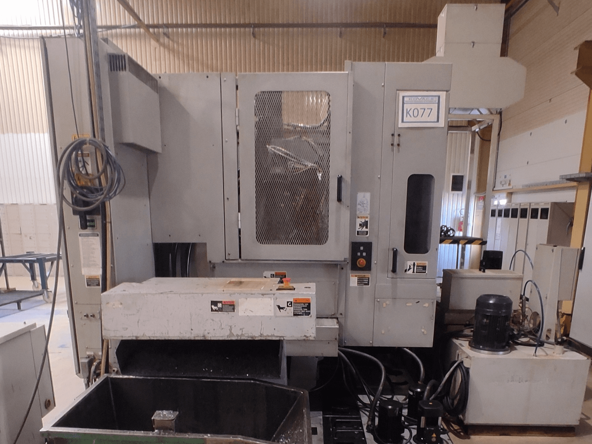 Máquina CNC industrial cinzenta com uma grande mesa de trabalho, vista lateral, com painel de controlo e reservatório de líquido de refrigeração, modelo K077.