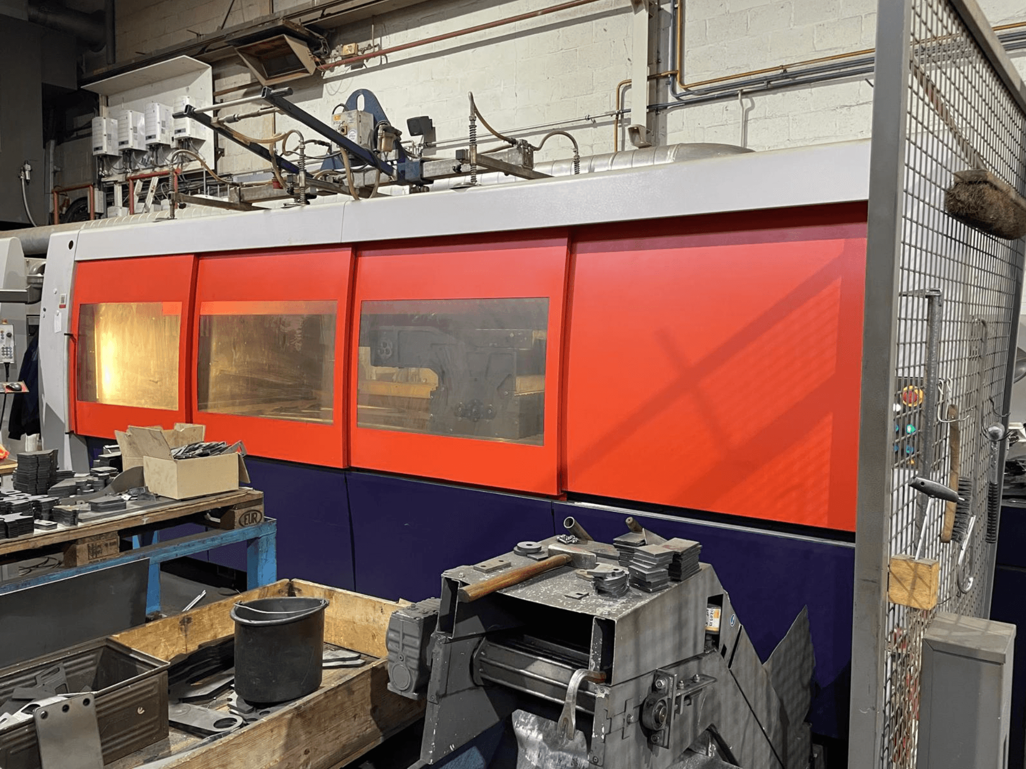 Grande plano de uma máquina de corte a laser industrial laranja e branca, vista lateral, com painel de controlo e ferramentas de oficina em primeiro plano.