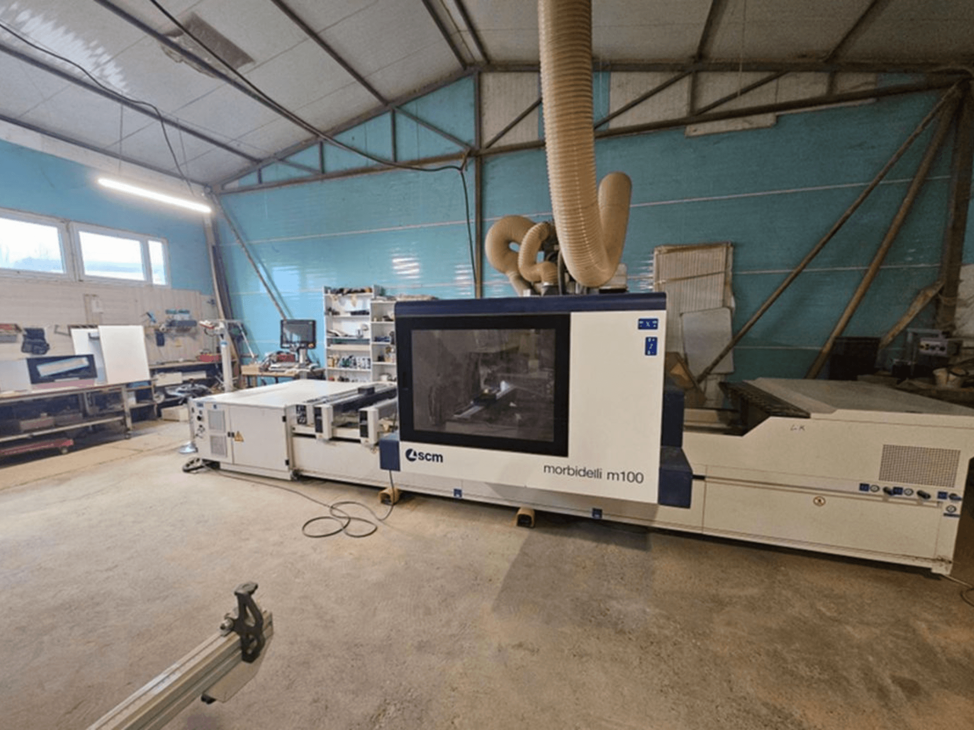 Centro de usinagem CNC SCM MORBIDELLI M 100 numa oficina, vista frontal com sistema de extração de poeiras e equipamento do espaço de trabalho circundante.