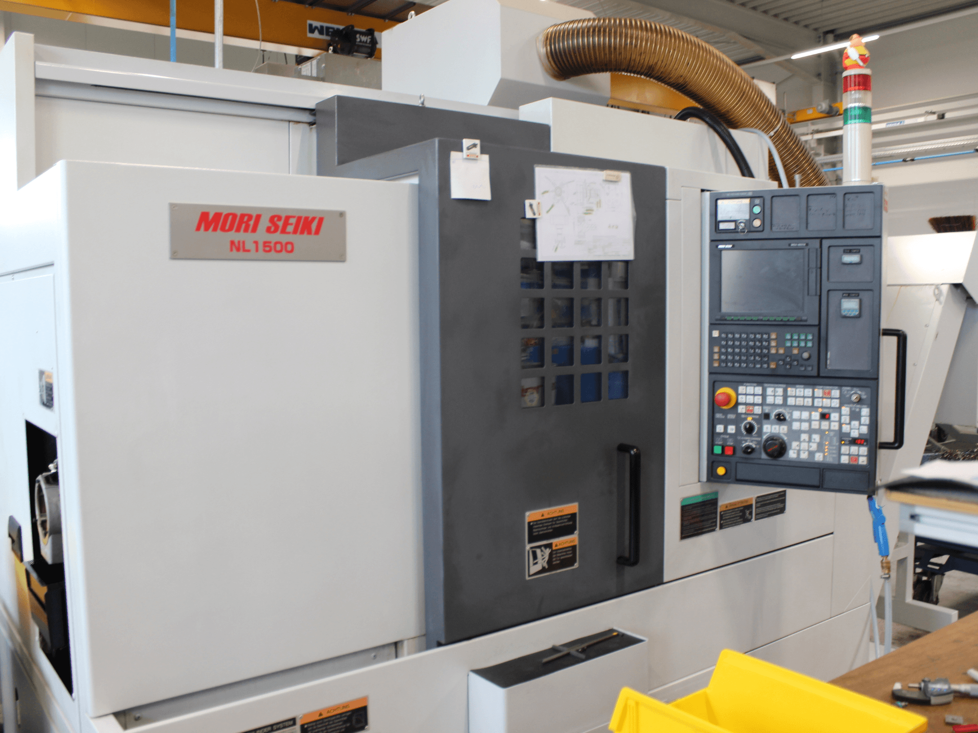 Centro de torneamento e fresagem MORI SEIKI NL 1500 SY, visto de lado, apresentando o painel de controlo, as etiquetas de segurança operacional e o sistema de arrefecimento.