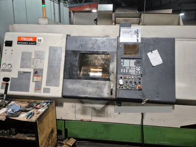 Torno Mazak Integrex 200Y, vista frontal, com painel de controlo e ferramentas sobre uma bancada. Indicadores visíveis e ecrã de visualização.